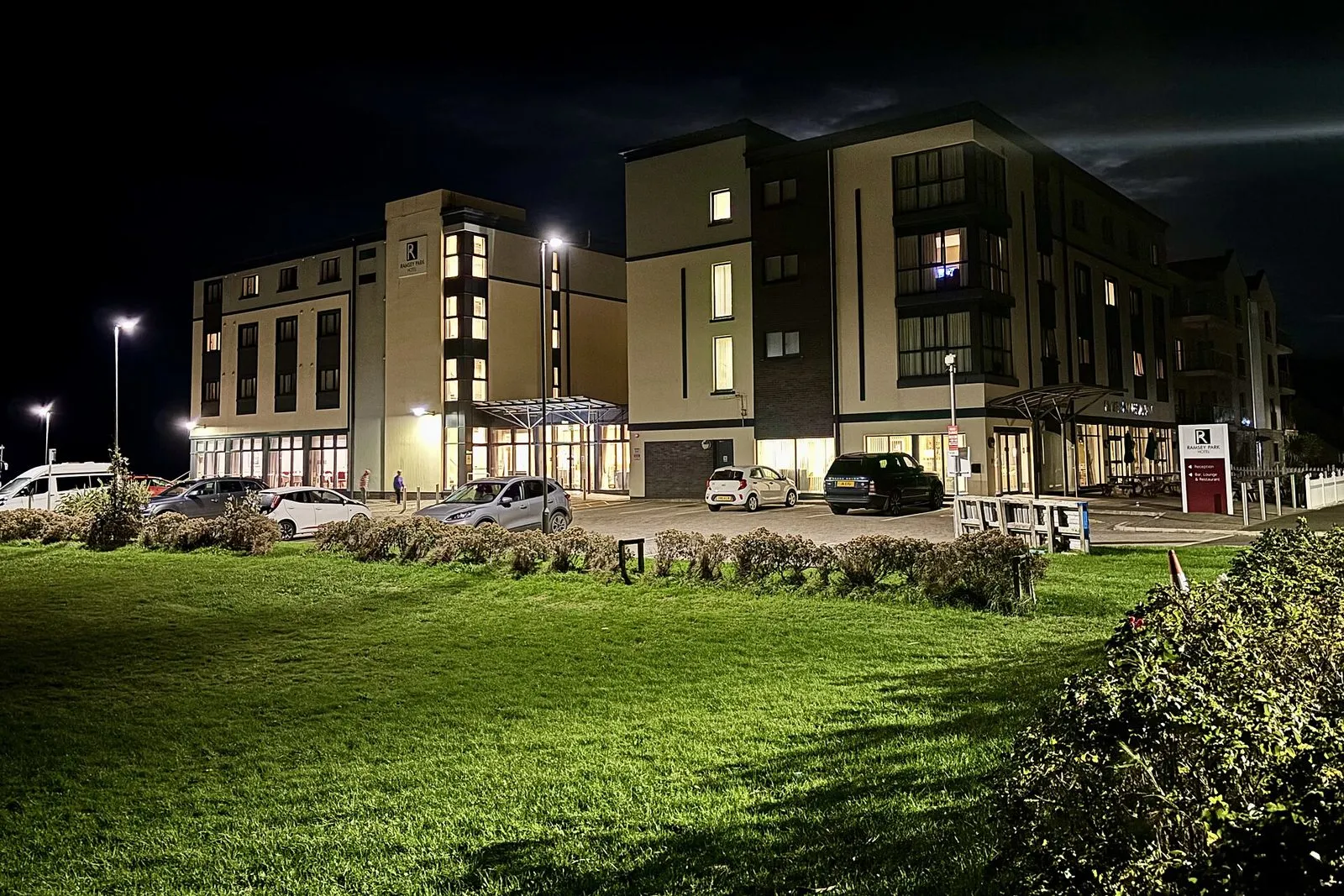 A modern four-storey hotel lit up at night beside a lawn, with cars parked out front