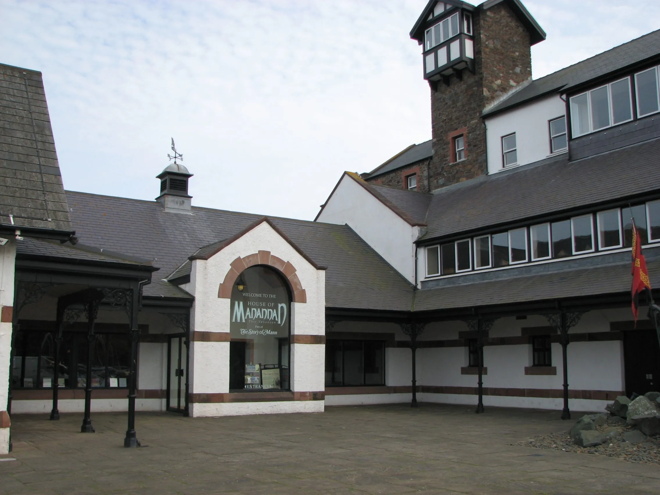 The white-walled courtyard entrance of the House of Manannan in Peel, with its arched sign, slate roofs and a stone tower, in the old railway station buildings