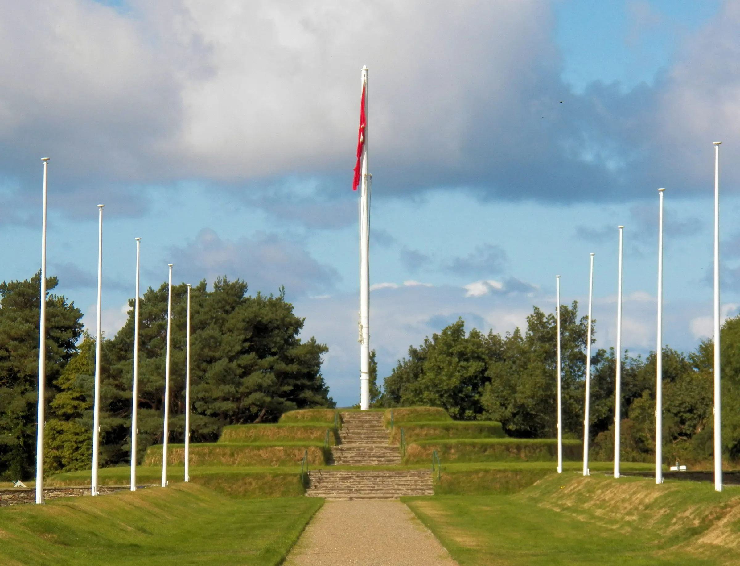 Tynwald Hill at St John's, a stepped grass mound with a path leading up to a flagpole flying the red Manx flag, lined with white flagpoles