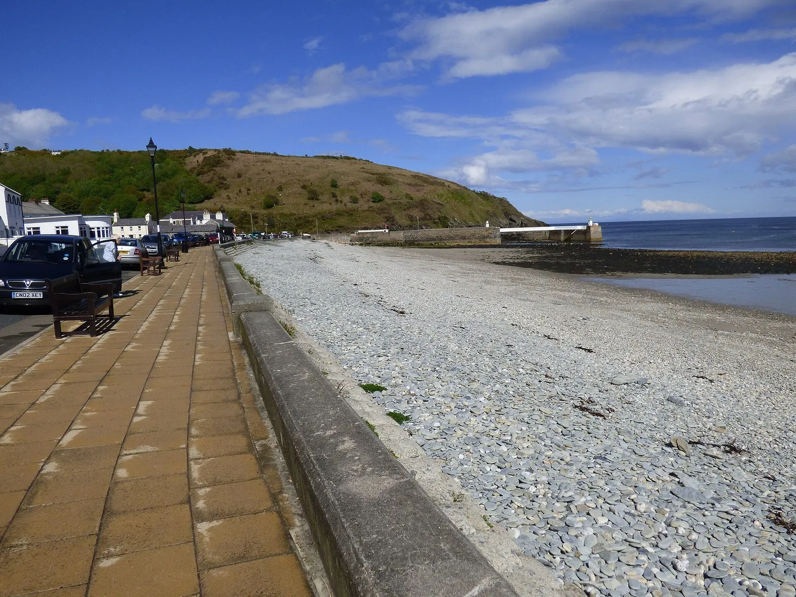 A wide stony beach curving to the harbour wall and a small lighthouse at Old Laxey, with a paved promenade and parked cars on the left and a green headland behind
