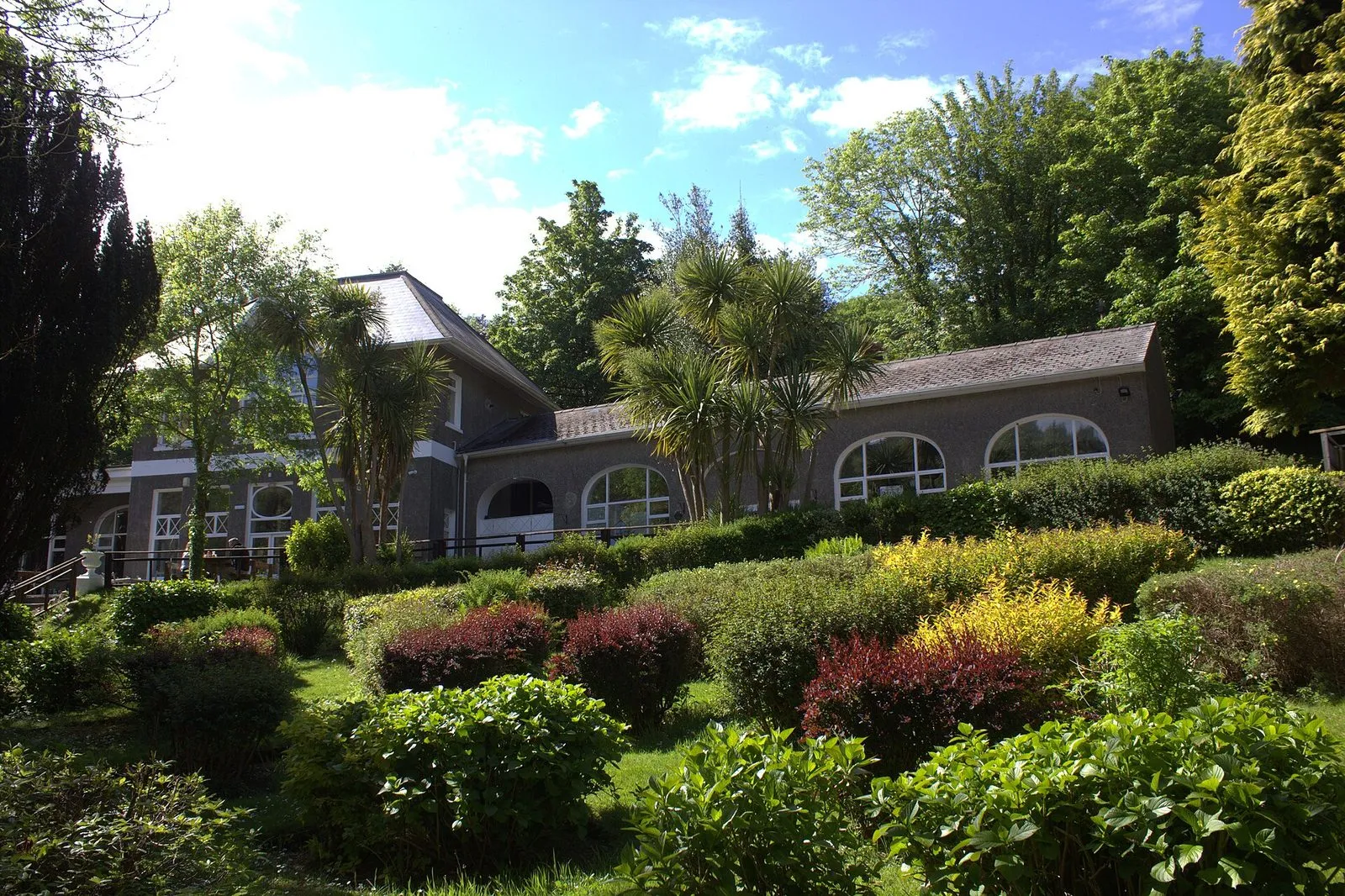 A grey-rendered Victorian pavilion with arched windows, framed by palms and flowering shrubs in Laxey Glen Gardens