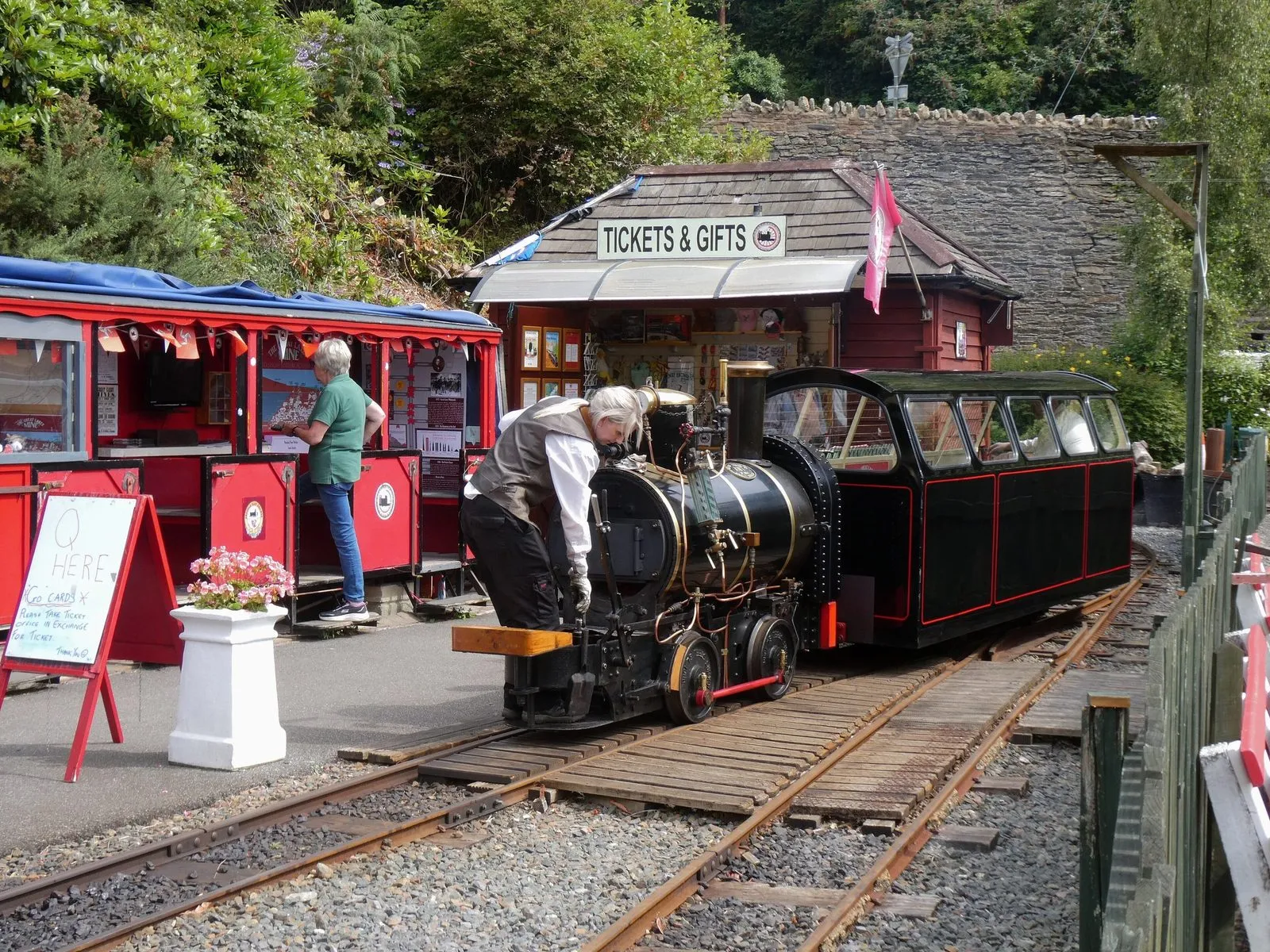 A small black steam locomotive at the Great Laxey Mine Railway station, with a volunteer driver oiling it beside the ticket office