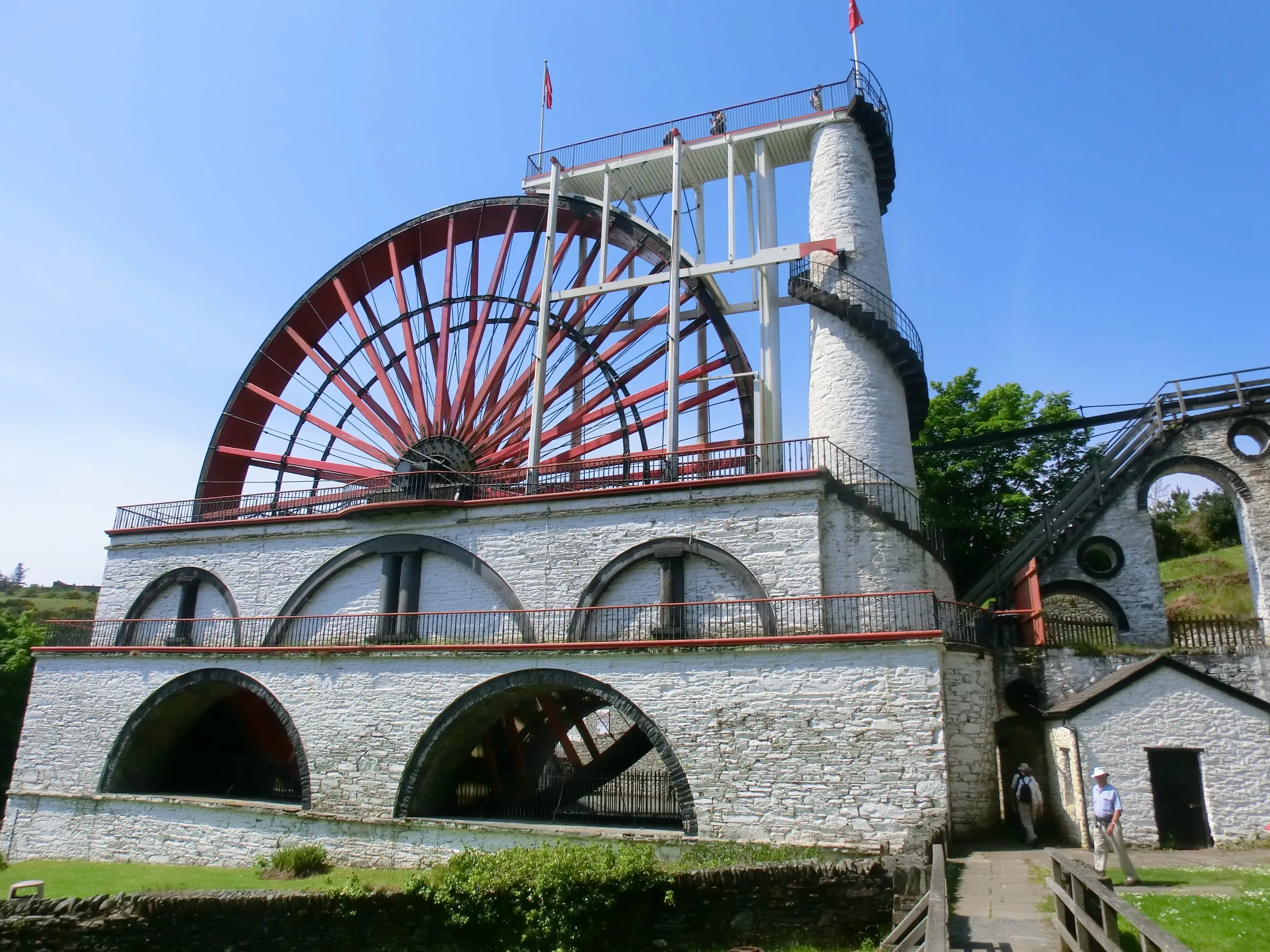 The Great Laxey Wheel, its red spokes above the white stone wheelcase and the spiral staircase tower, under a clear blue sky