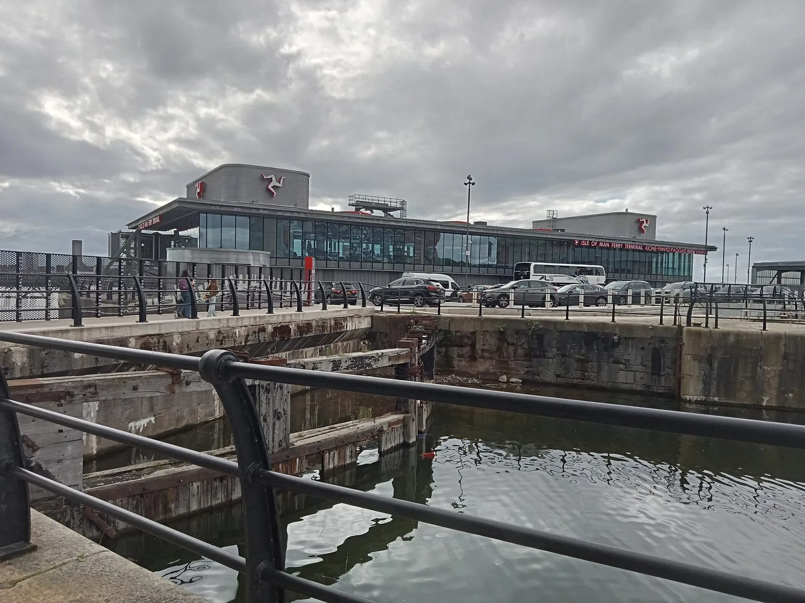 The Liverpool ferry terminal seen across an old dock basin, with cars parked in front of its long glass frontage and the three-legs emblem on its roof