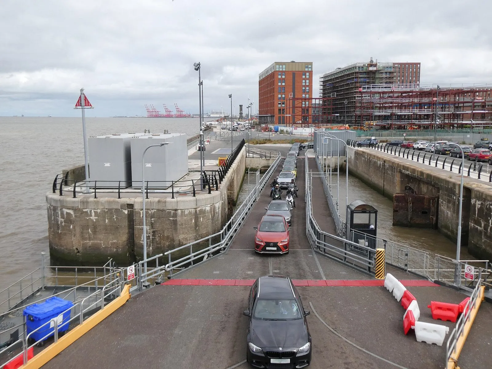 Cars driving off the ramp from a ferry onto the quay at the Liverpool terminal, with the Mersey on the left and new flats going up on the right
