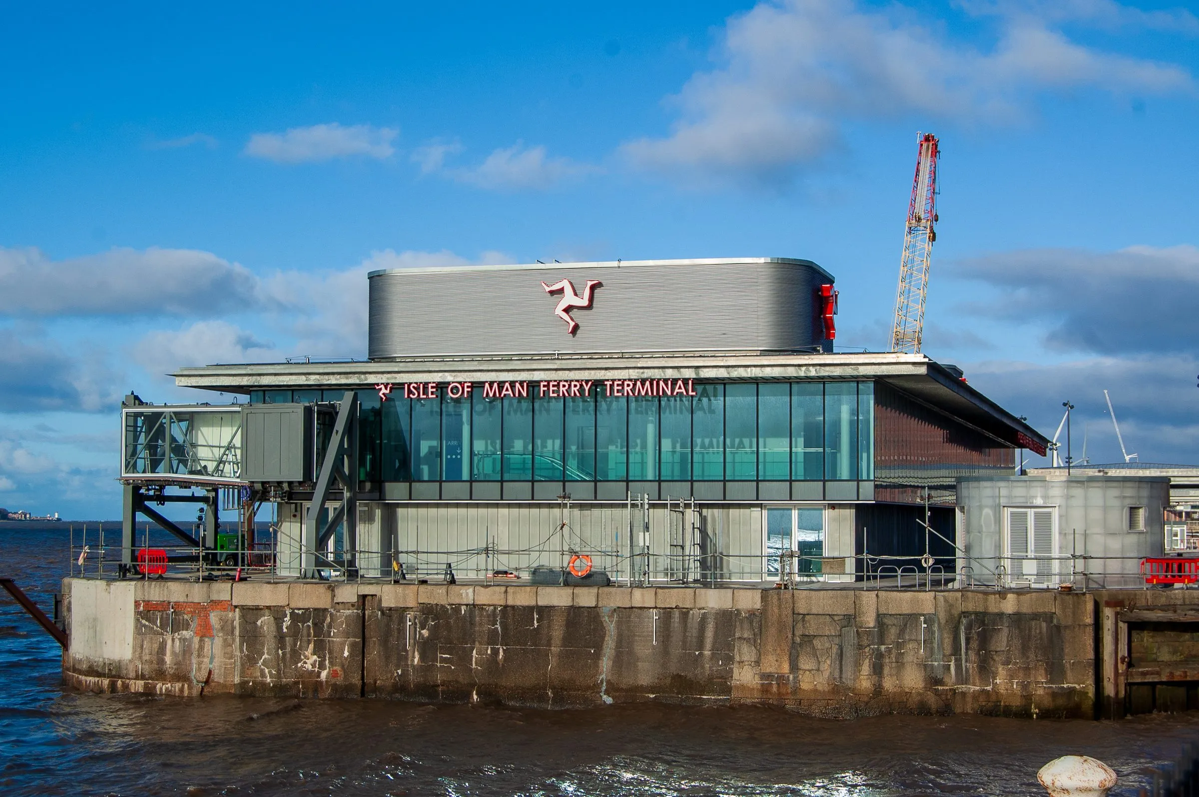 The Isle of Man Ferry Terminal in Liverpool, a glass and grey-metal building with a large three-legs emblem, on the quay beside the Mersey under a blue sky