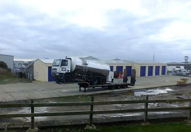 A white milk tanker lorry parked on a low-loader outside a row of blue-doored industrial units under a grey sky