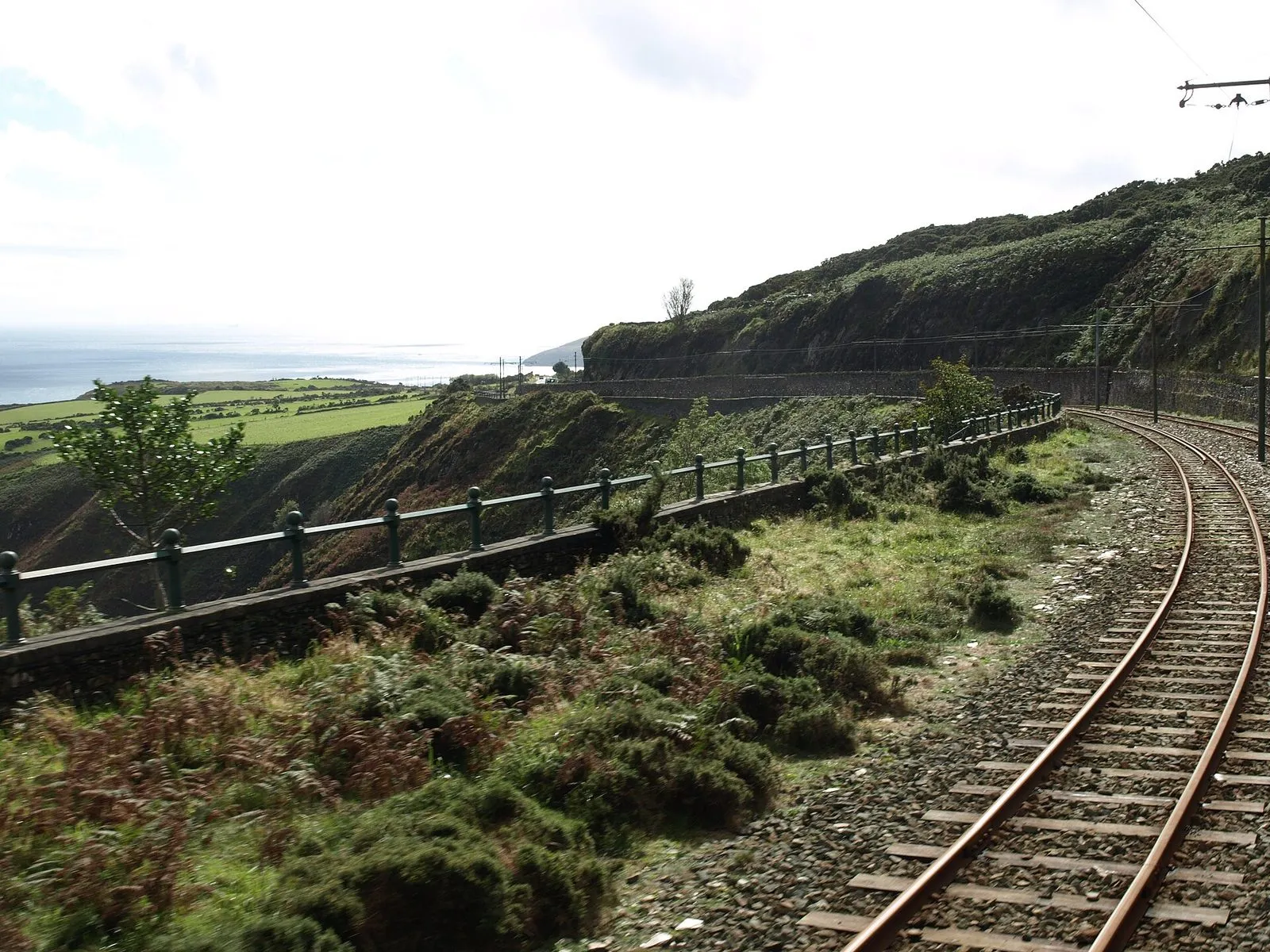 A curving tram line on a walled ledge above Bulgham Bay, with green fields and the sea below
