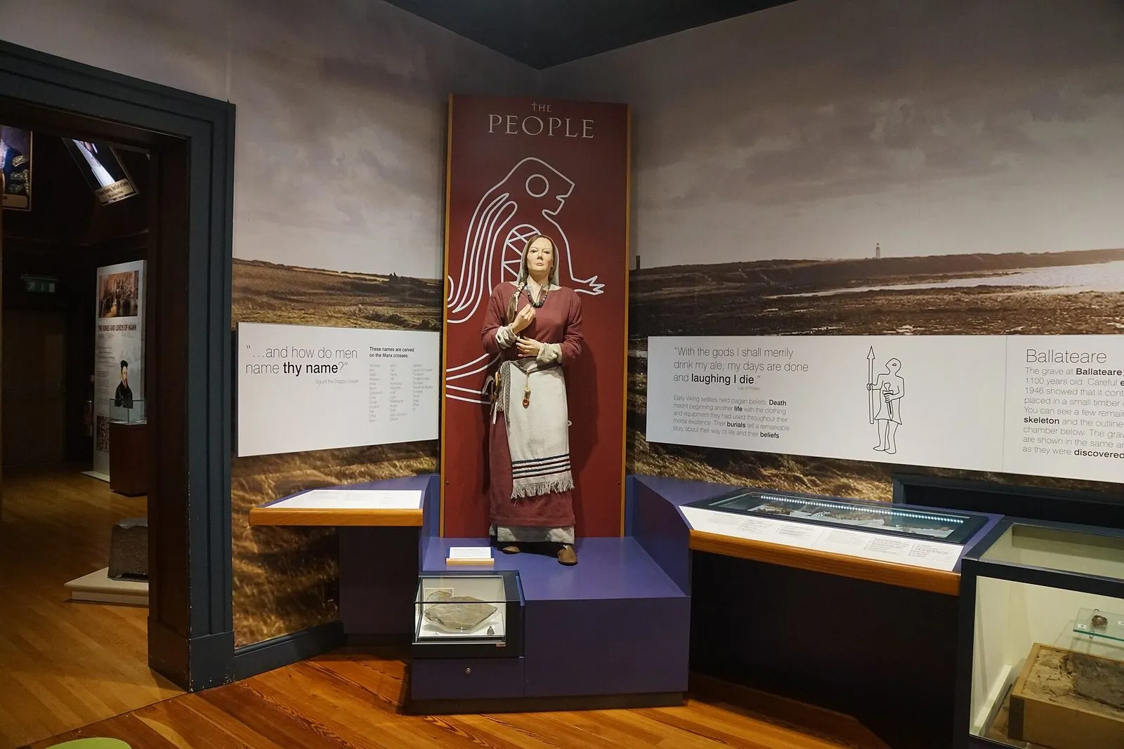 A lifelike figure of a Viking-age woman in a red dress and white apron, displayed in the Manx Museum’s Viking gallery beside information panels about burials