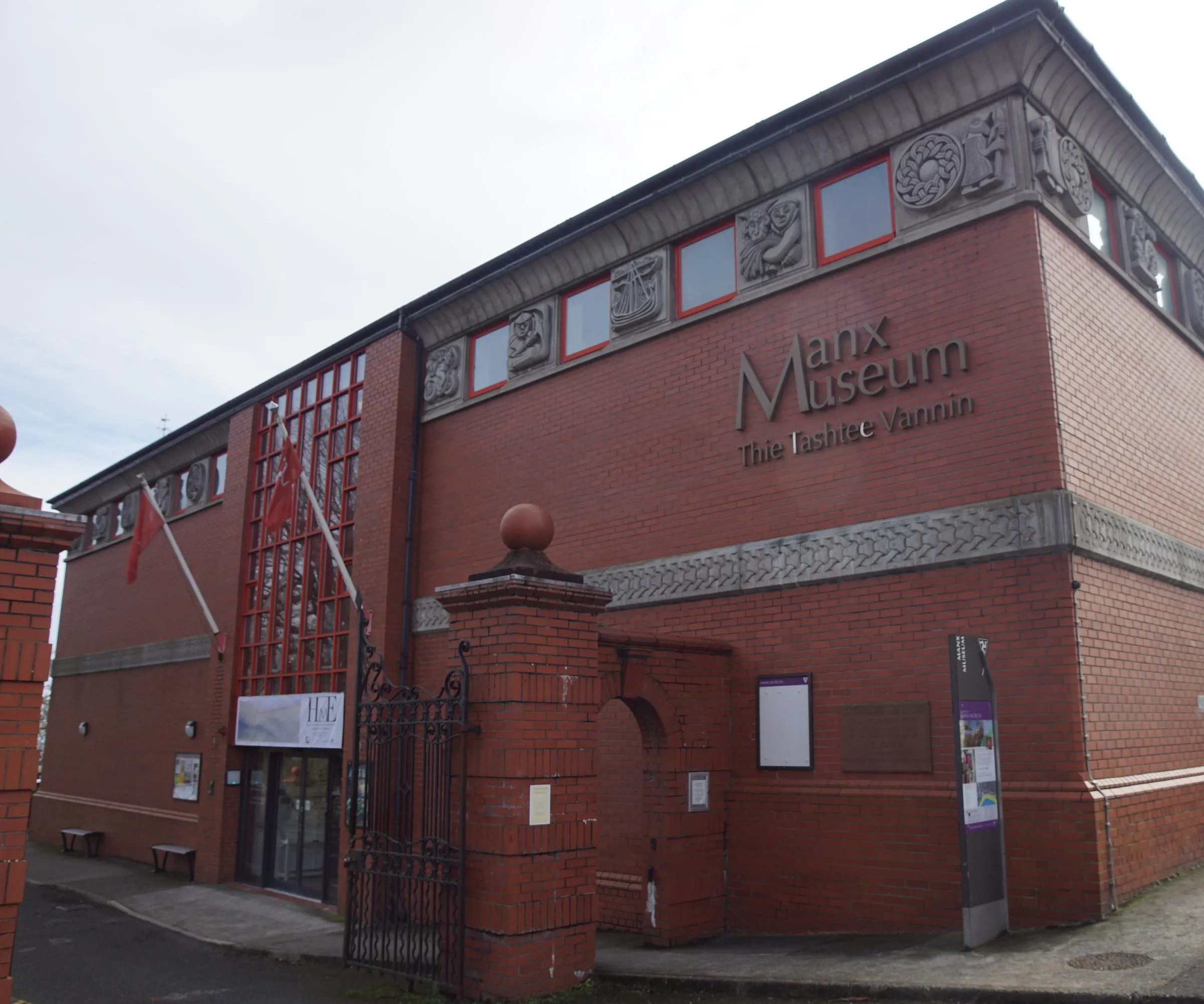 The red-brick front of the Manx Museum in Douglas, with carved stone panels under the roofline, the gates and the Manx flag by the entrance