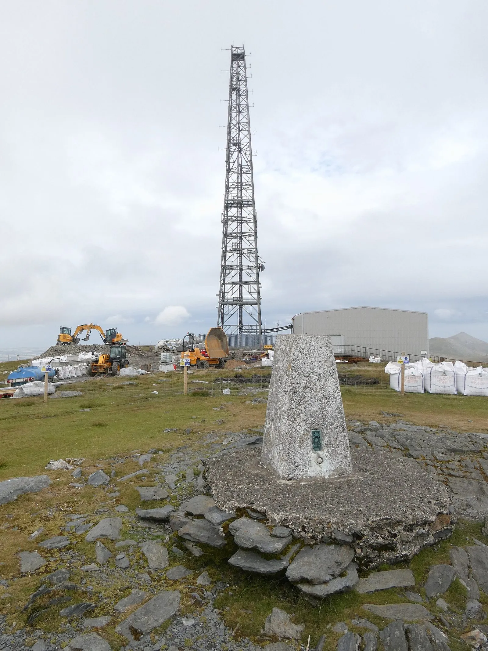 A tall steel lattice radio mast beside the concrete trig point on the summit of Snaefell, with construction machinery and a plant building nearby