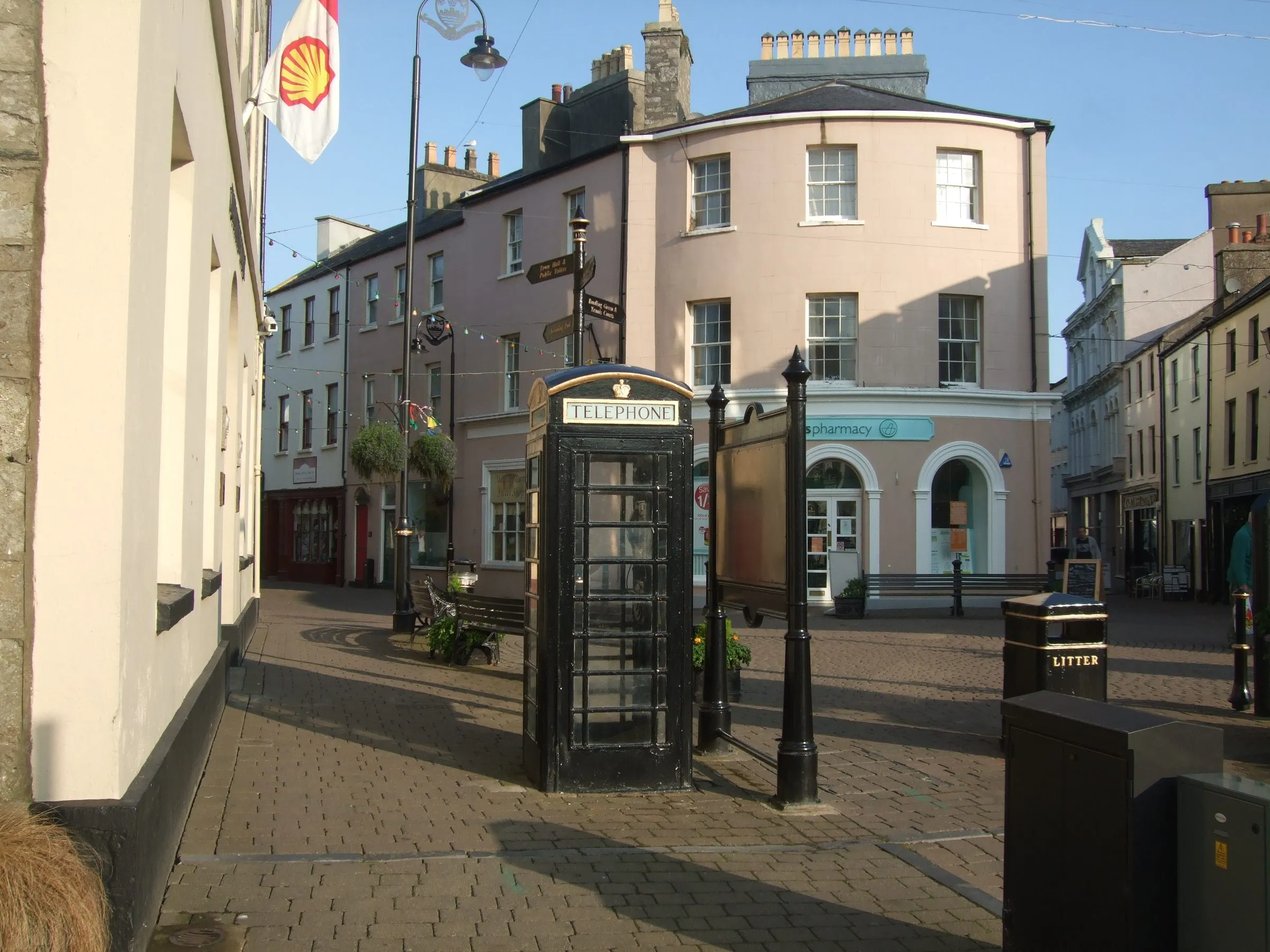 A black cast-iron telephone box in Castletown Square, in front of pastel-painted town-centre buildings on a sunny evening