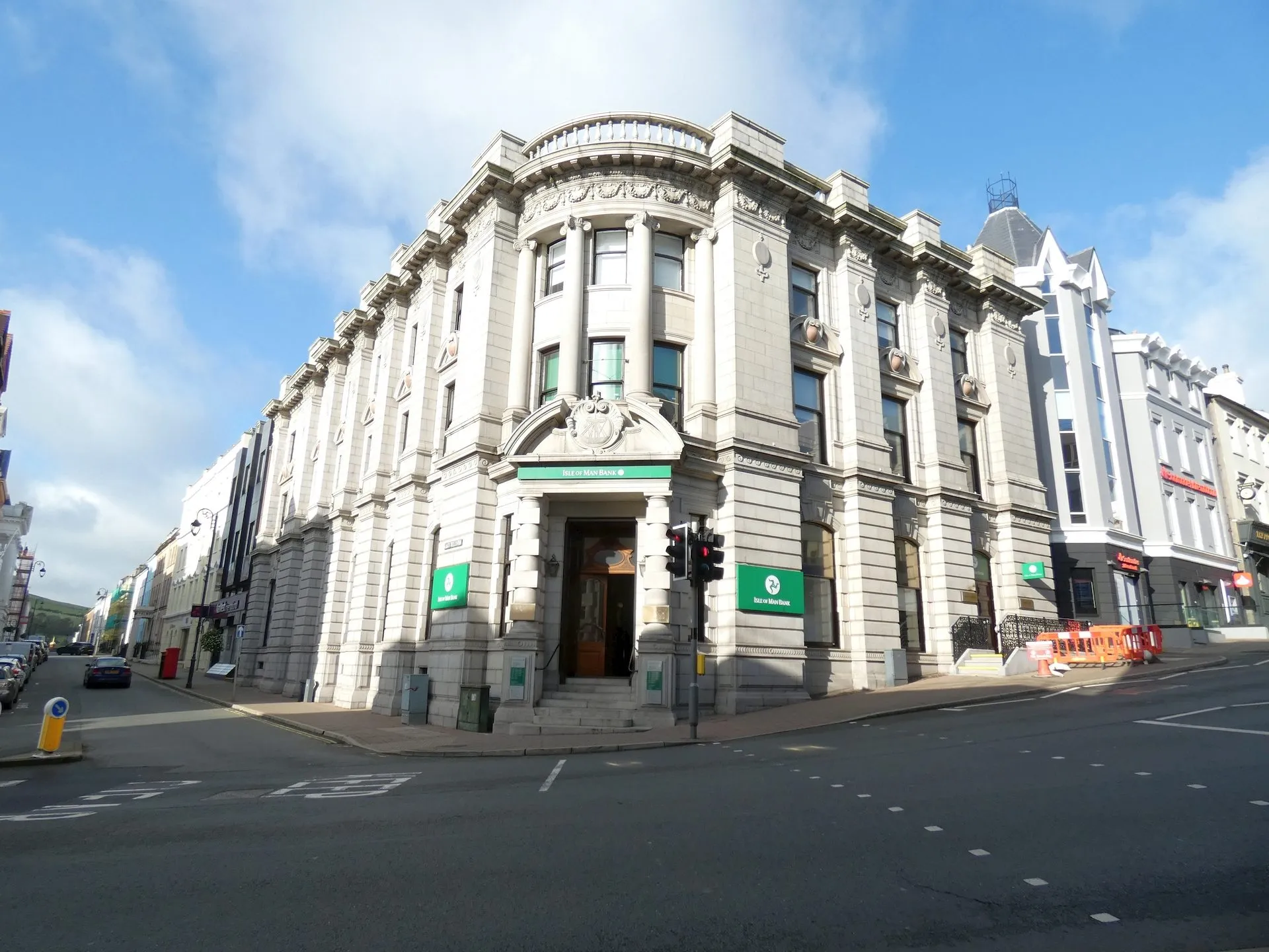 The grand granite corner building of the Isle of Man Bank in Douglas under a blue sky, with green bank signs by the door