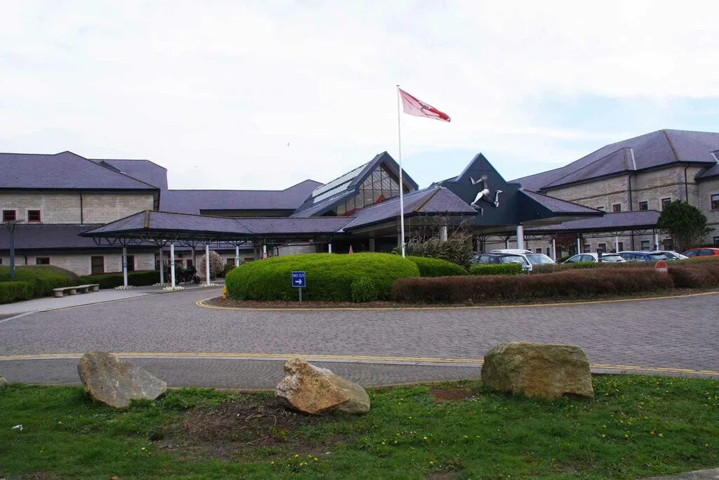 Noble’s Hospital seen across a paved turning circle, a long, low stone building with slate roofs, a glazed gable and a Manx flag outside the main entrance
