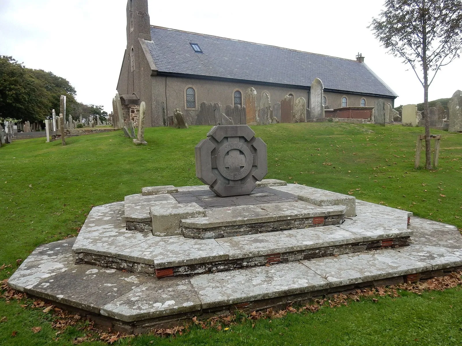 A long grey stone church with arched windows behind a graveyard of old headstones and an octagonal stone memorial on steps in the foreground