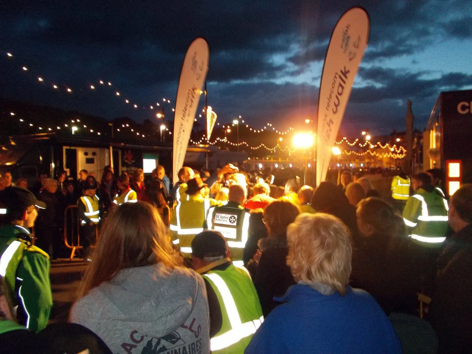 A crowd in hi-vis jackets under festoon lights and Parish Walk banners at the finish on Douglas promenade at dusk