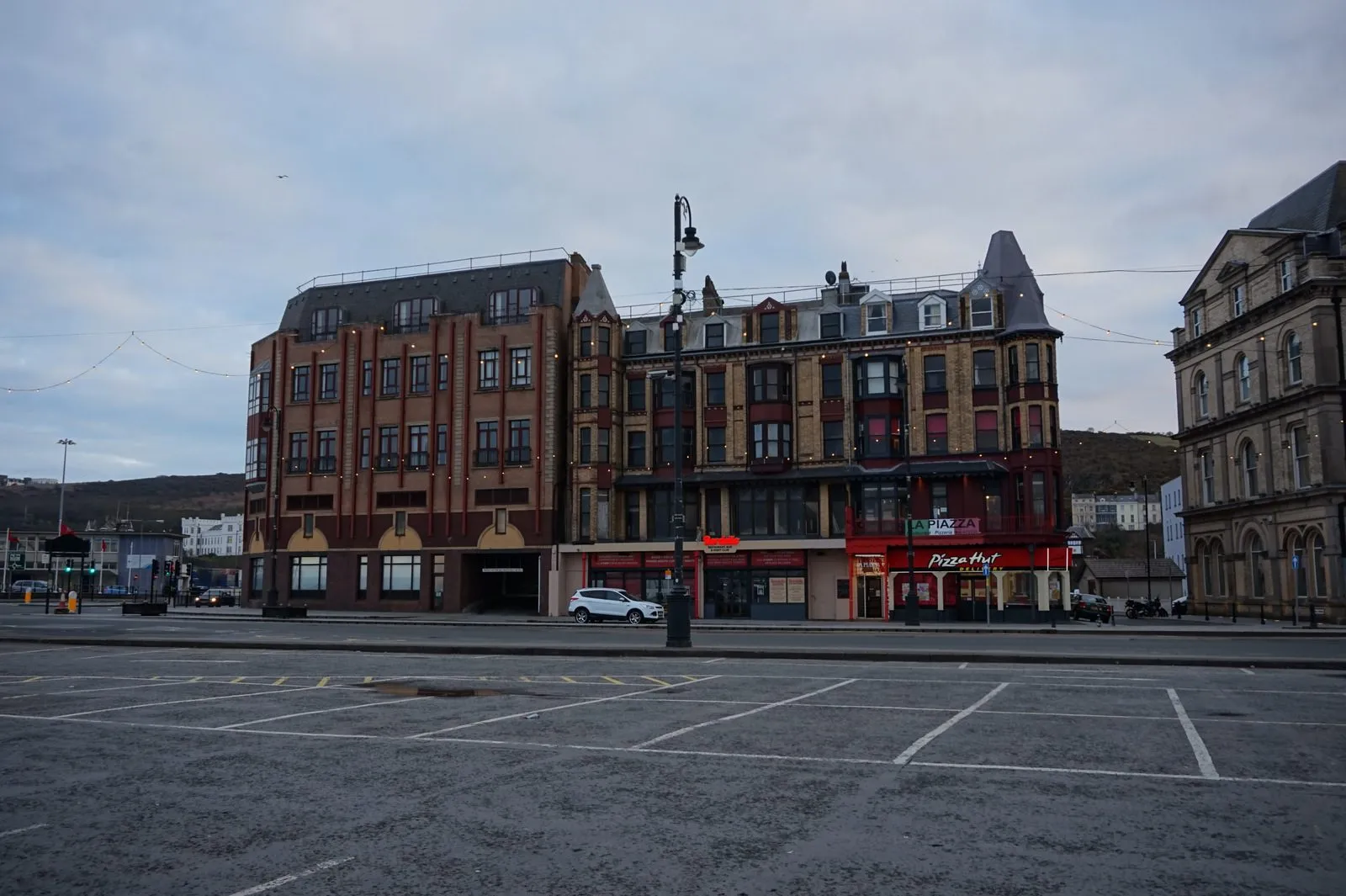 A marked-out car park on Loch Promenade in Douglas, with red-brick and stone buildings behind it under a grey evening sky