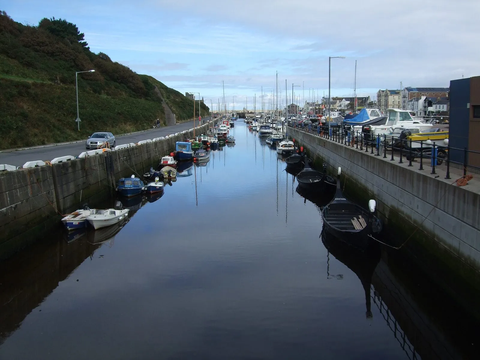 Peel marina on a still morning, small boats moored along both walls of the narrow harbour with the green slope of Peel Hill on the left