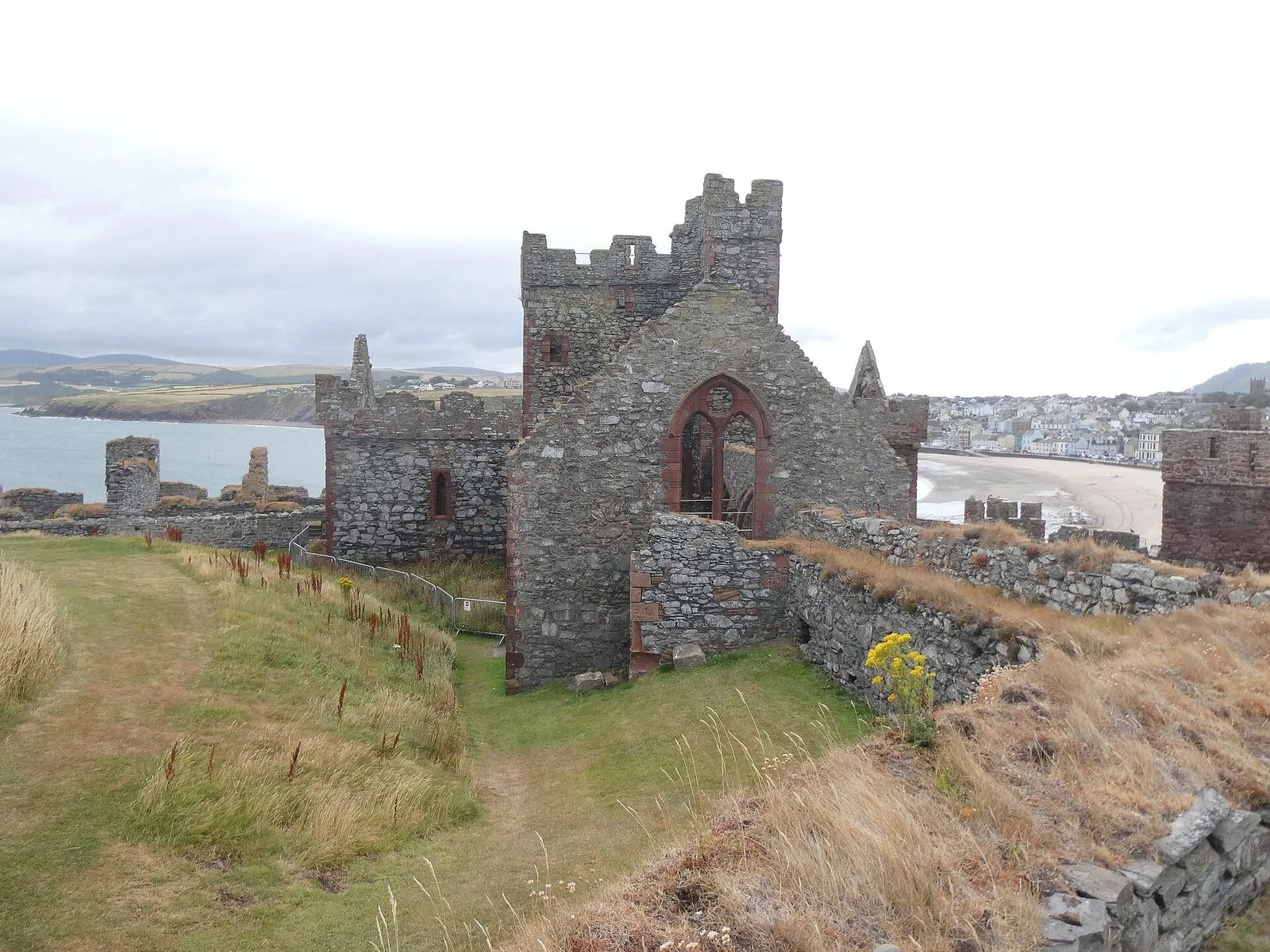 The ruined gable of St German’s Cathedral, with a pointed red sandstone window, rising above the grassy paths inside Peel Castle, with Peel beach behind
