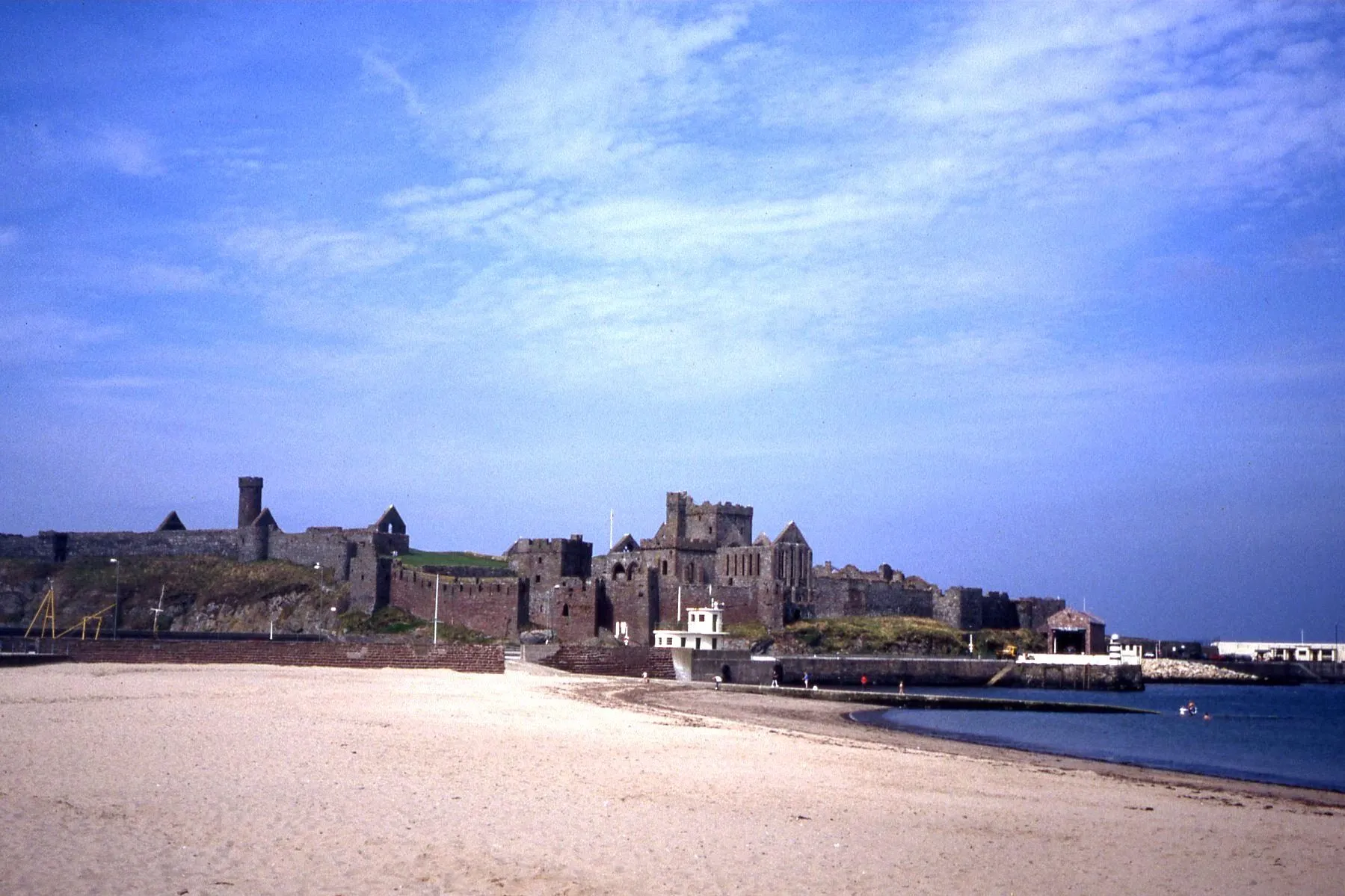 The red sandstone walls, round tower and ruined cathedral of Peel Castle on St Patrick's Isle, seen across the sand of Peel beach under a blue sky
