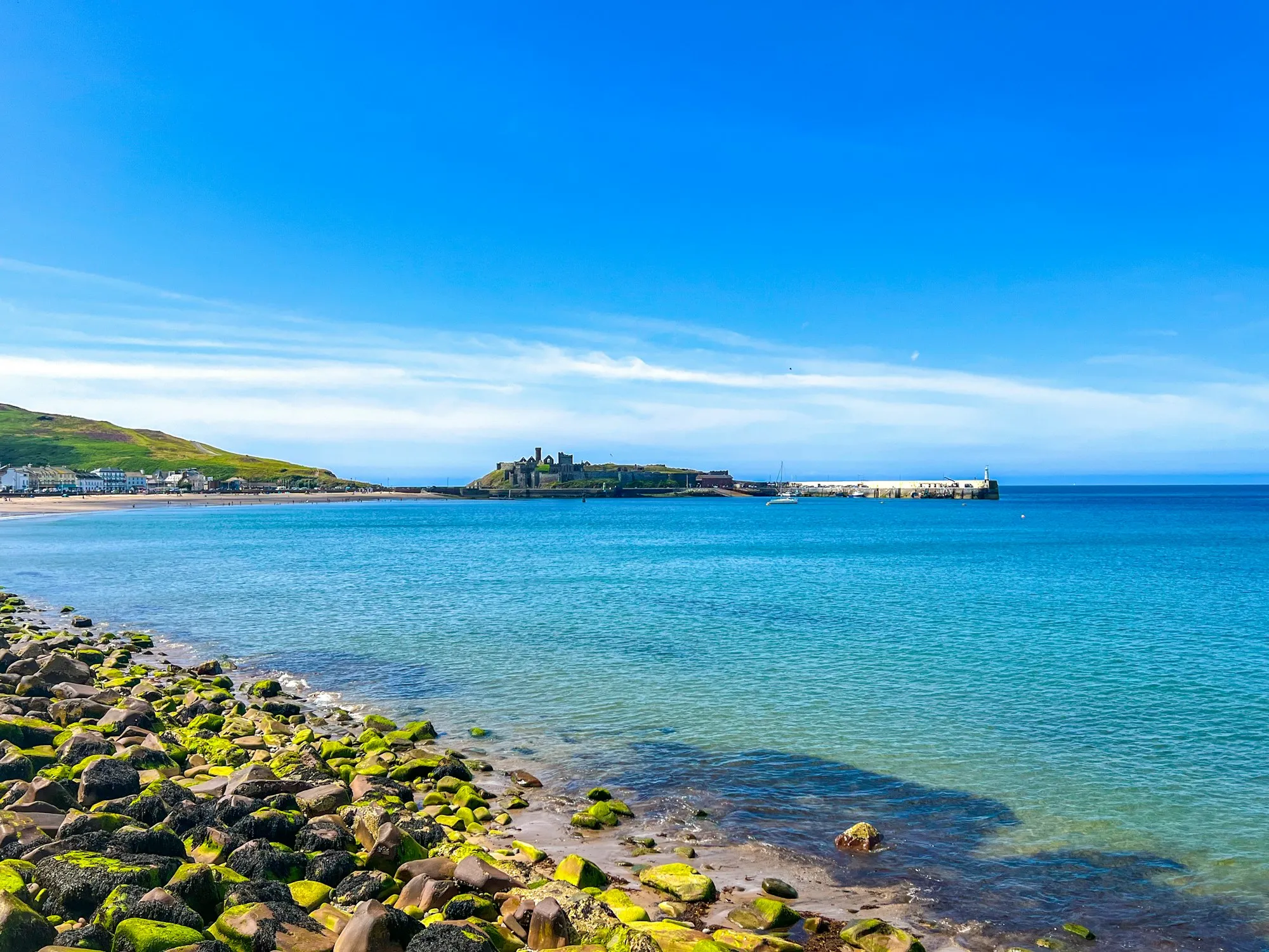 Peel Castle on St Patrick's Isle and the harbour breakwater, seen across the bay from the rocks on a bright blue day