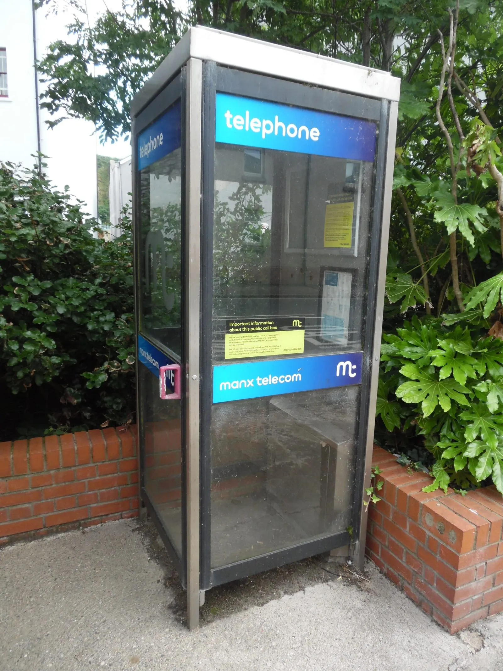 A modern glass Manx Telecom telephone box with blue “telephone” signs, between a low brick wall and green shrubs