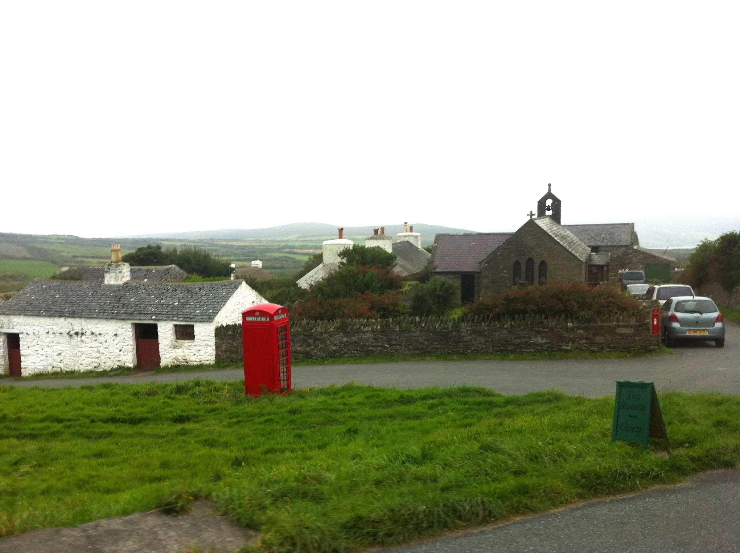 A red telephone box on the grass verge in Cregneash, with whitewashed cottages, a small stone church and green hills behind under a grey sky