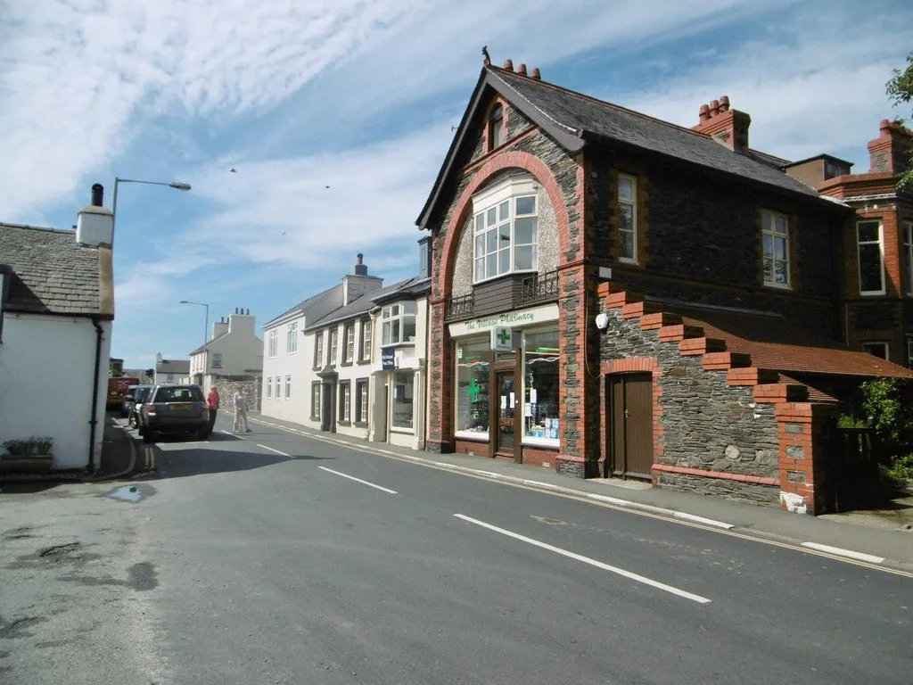 The Village Pharmacy in Kirk Michael, a red-sandstone and slate building with a green cross over the shop window, on the main road through the village