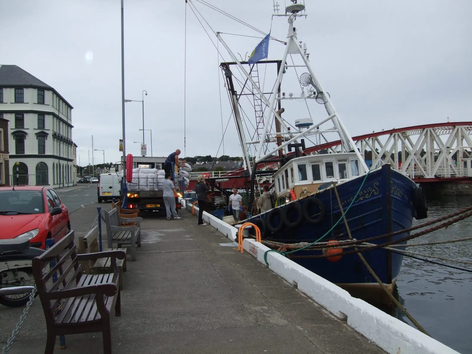A fishing boat moored against the quay in Ramsey harbour while sacks of queenies are loaded onto a lorry, with the swing bridge behind