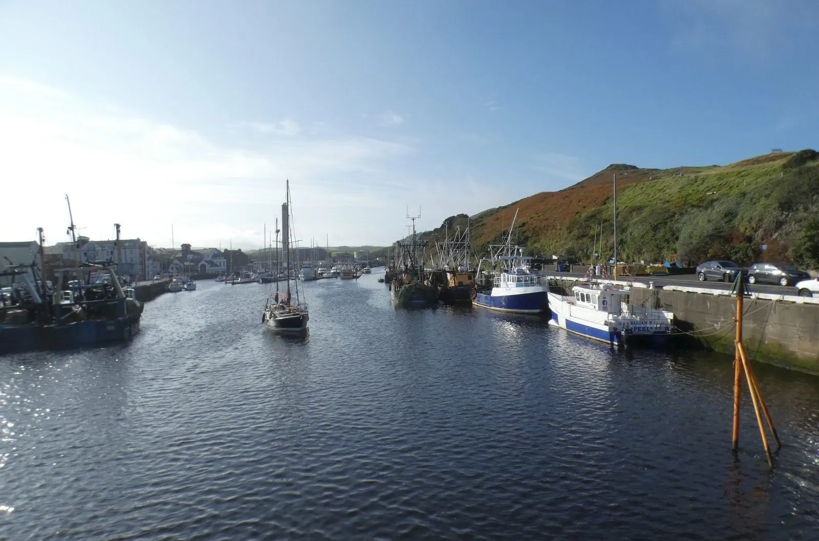 Fishing boats and yachts moored in Peel’s inner harbour under a green hillside on a bright day