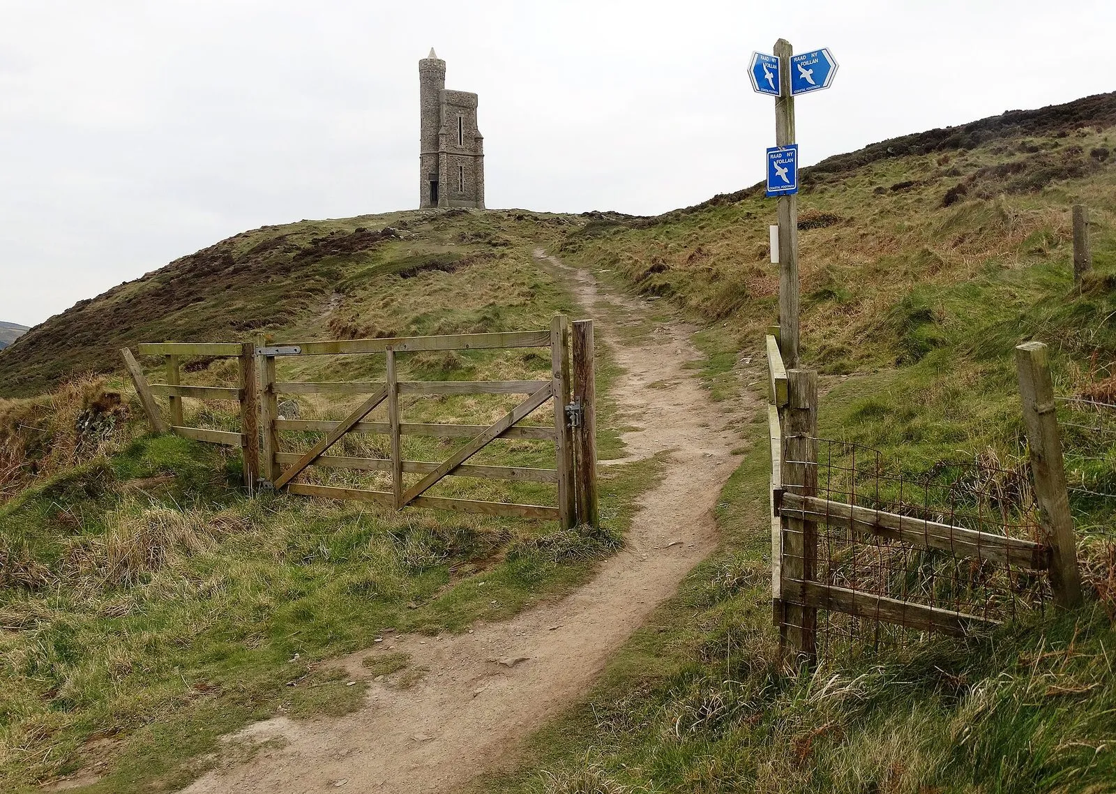 A worn dirt path leading through a wooden gate towards a stone tower on a hilltop, with a post carrying blue Raad ny Foillan gull waymarks