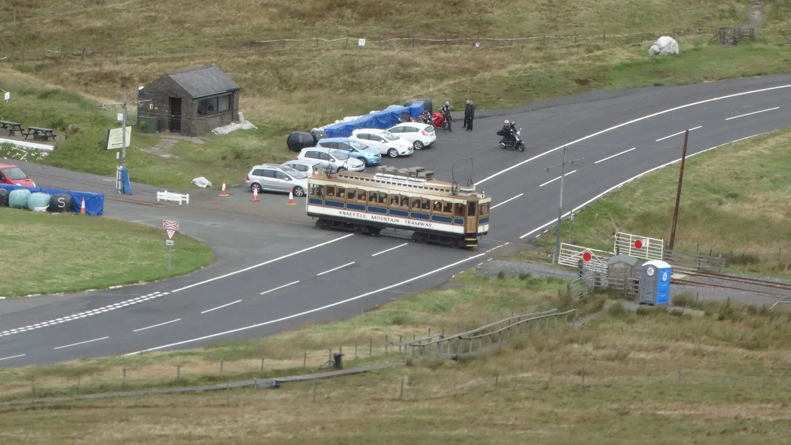 A Snaefell Mountain Railway tram crossing the empty Mountain Road at the Bungalow, with parked cars and motorbikes beside the road
