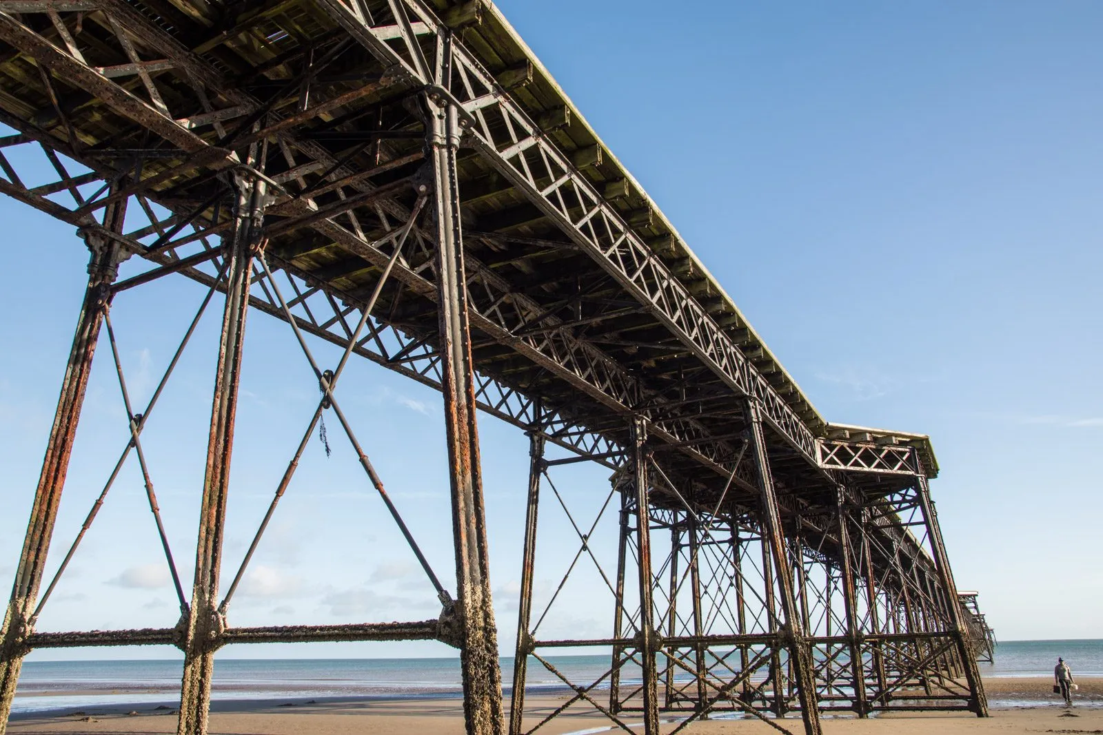 Looking up at the rusting iron legs and deck of Ramsey’s Queen’s Pier, stretching out across a sandy beach at low tide