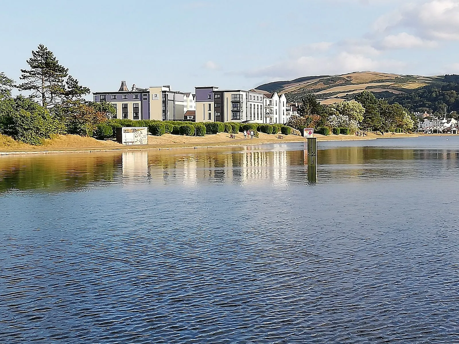 A calm lake in Mooragh Park with apartment blocks and trees along the far shore and the hills above Ramsey behind