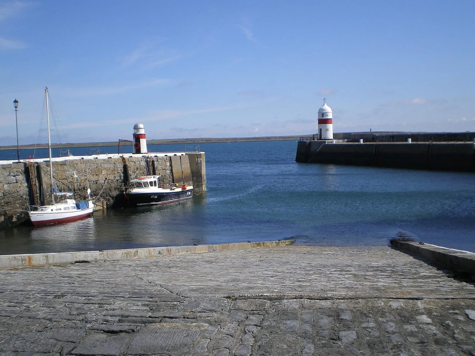 A cobbled slipway running down into Castletown harbour, with a fishing boat and a small yacht moored against the stone wall and two red-and-white harbour lights
