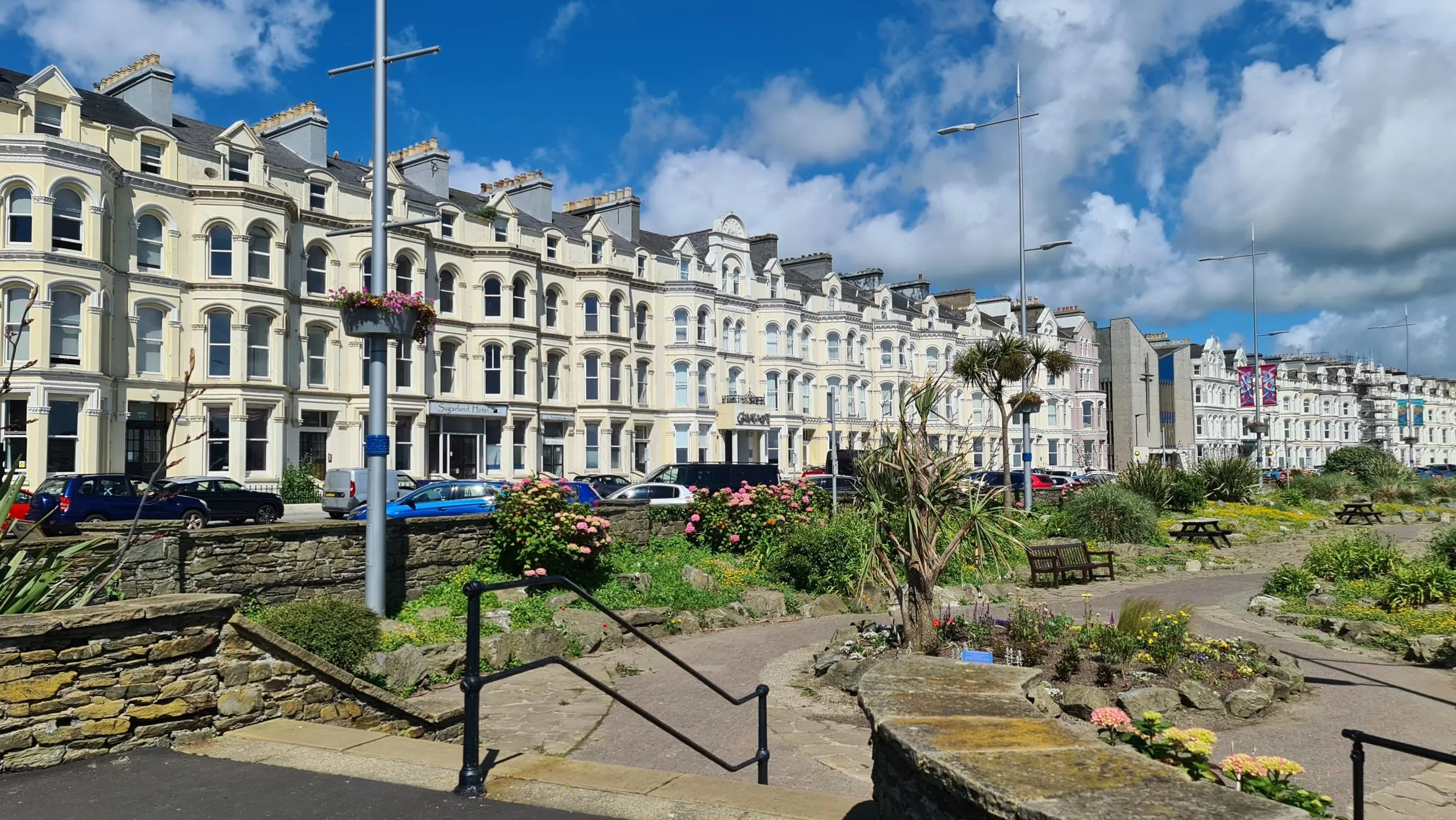 The cream Victorian hotel terraces of Loch Promenade in Douglas behind a sunken garden with palms and flower beds, under a blue sky