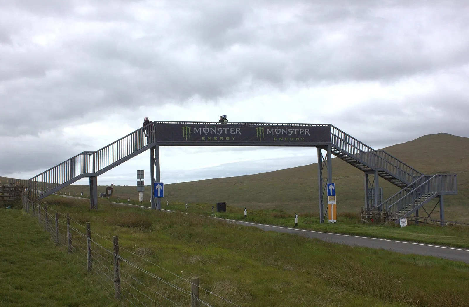 A steel footbridge over the Mountain Road at the Bungalow, with blue one-way arrow signs on the verge and bare moorland behind