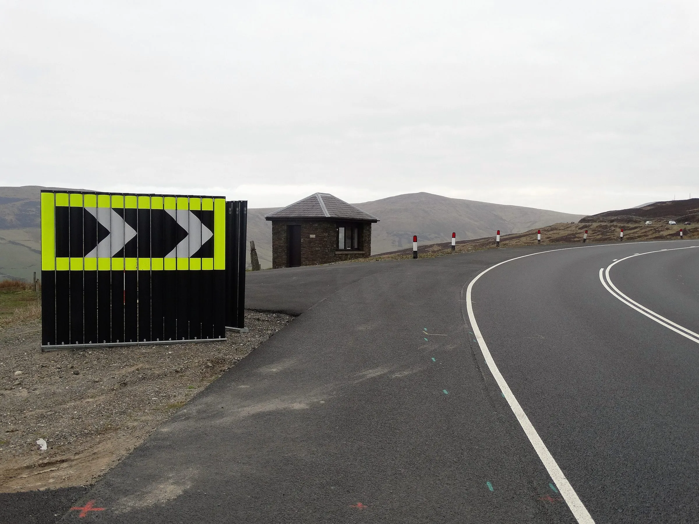 The open A18 Mountain Road curving right at Keppel Gate, with a black and yellow chevron board, a stone marshals' hut and moorland hills behind