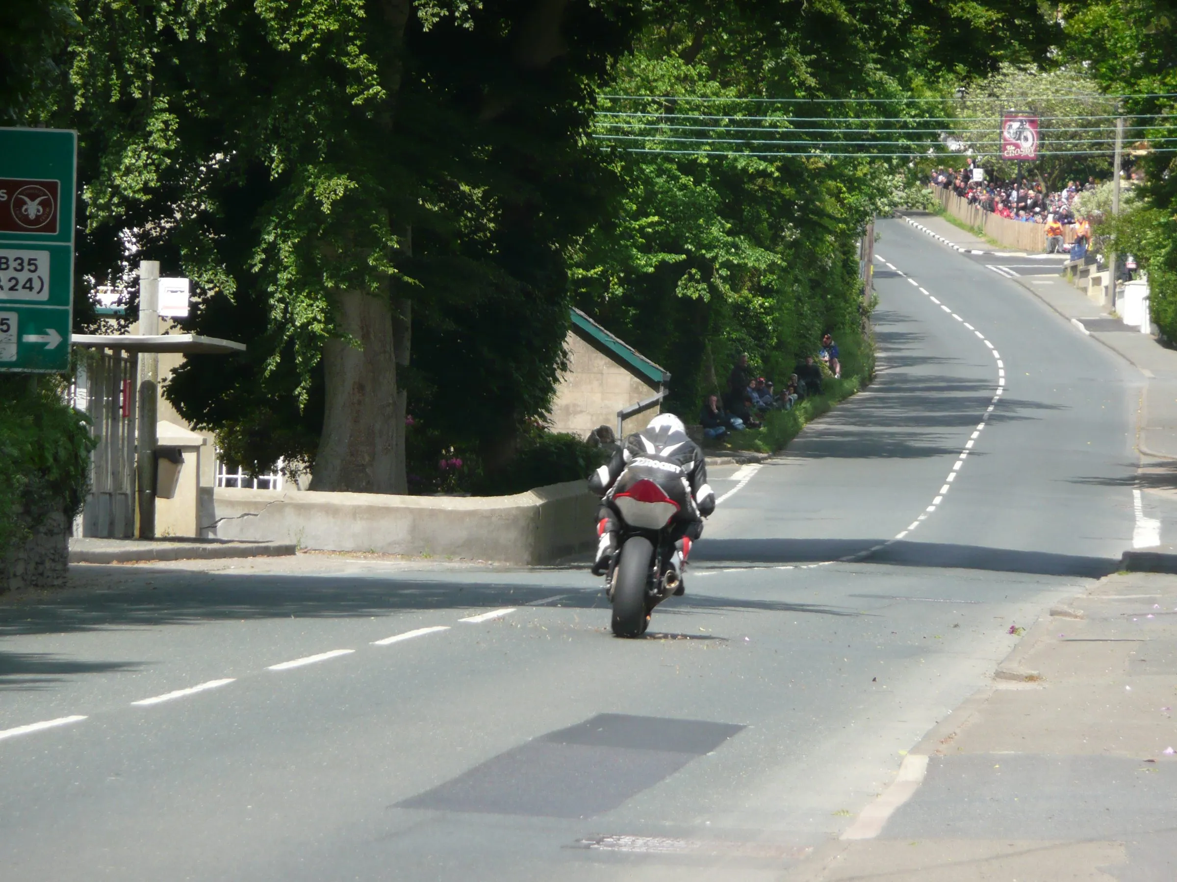 A racer riding away up the closed road towards Crosby crossroads, with spectators lining the bank and the verge behind the barriers