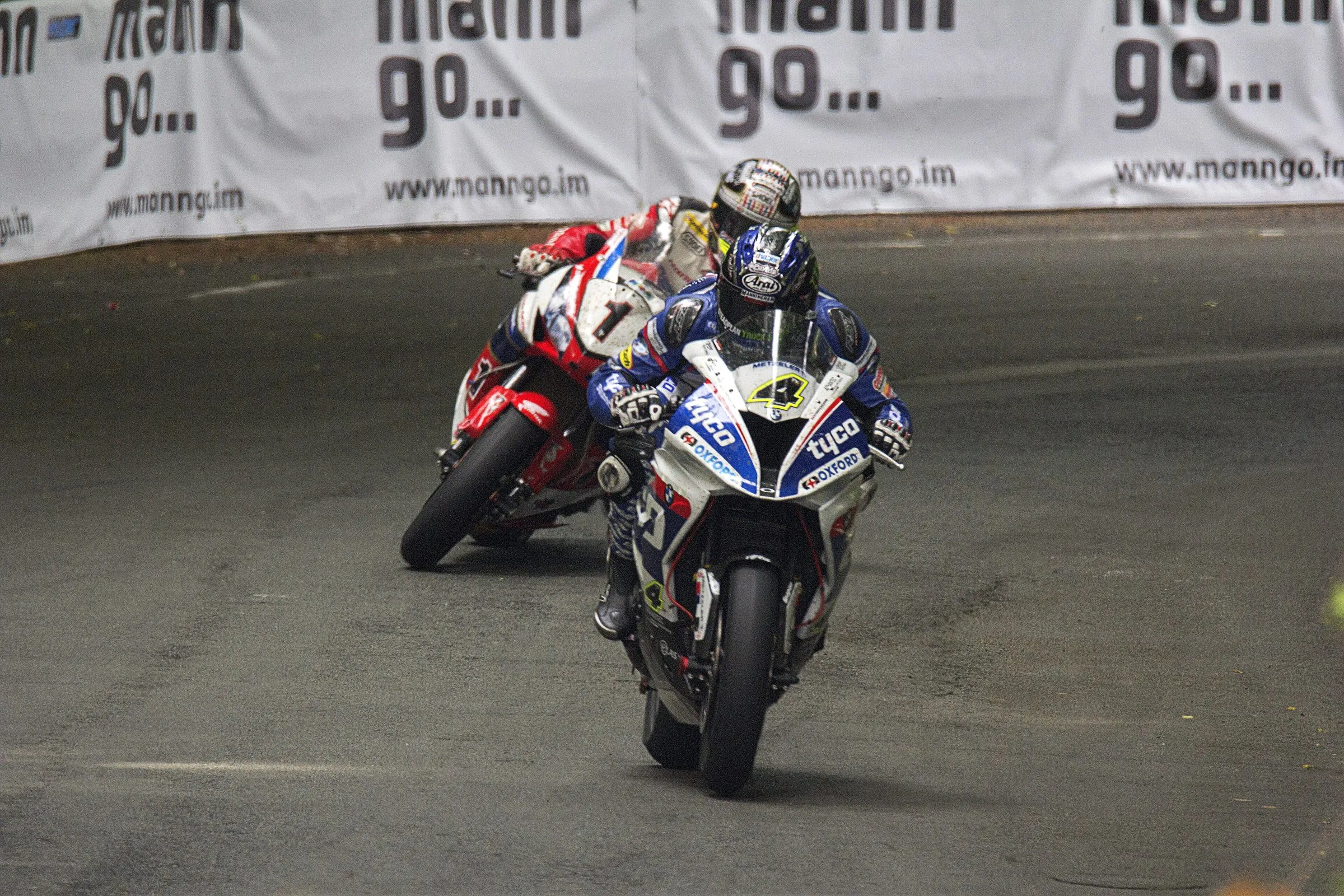 Two superbikes, numbers 4 and 1, racing nose to tail through a tree-shaded corner during the Senior TT