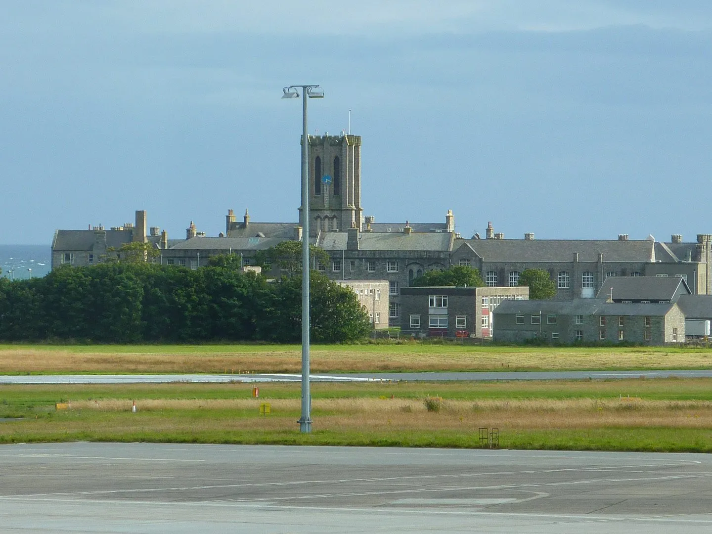 King William’s College, a grey stone Victorian building with a central tower, seen across the grass beside the airport runway