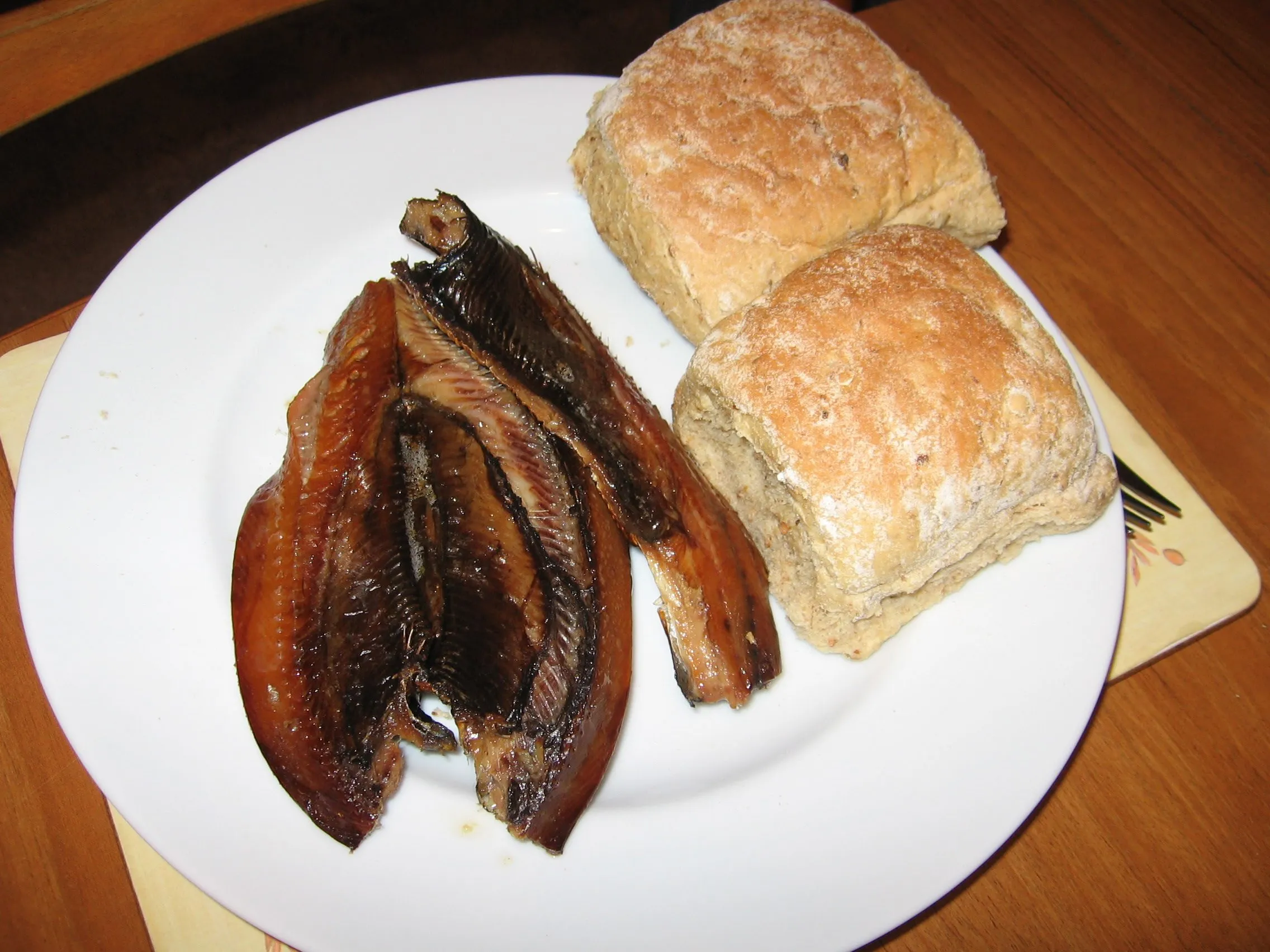 Two golden-brown smoked kipper fillets on a white plate beside two floury bread rolls