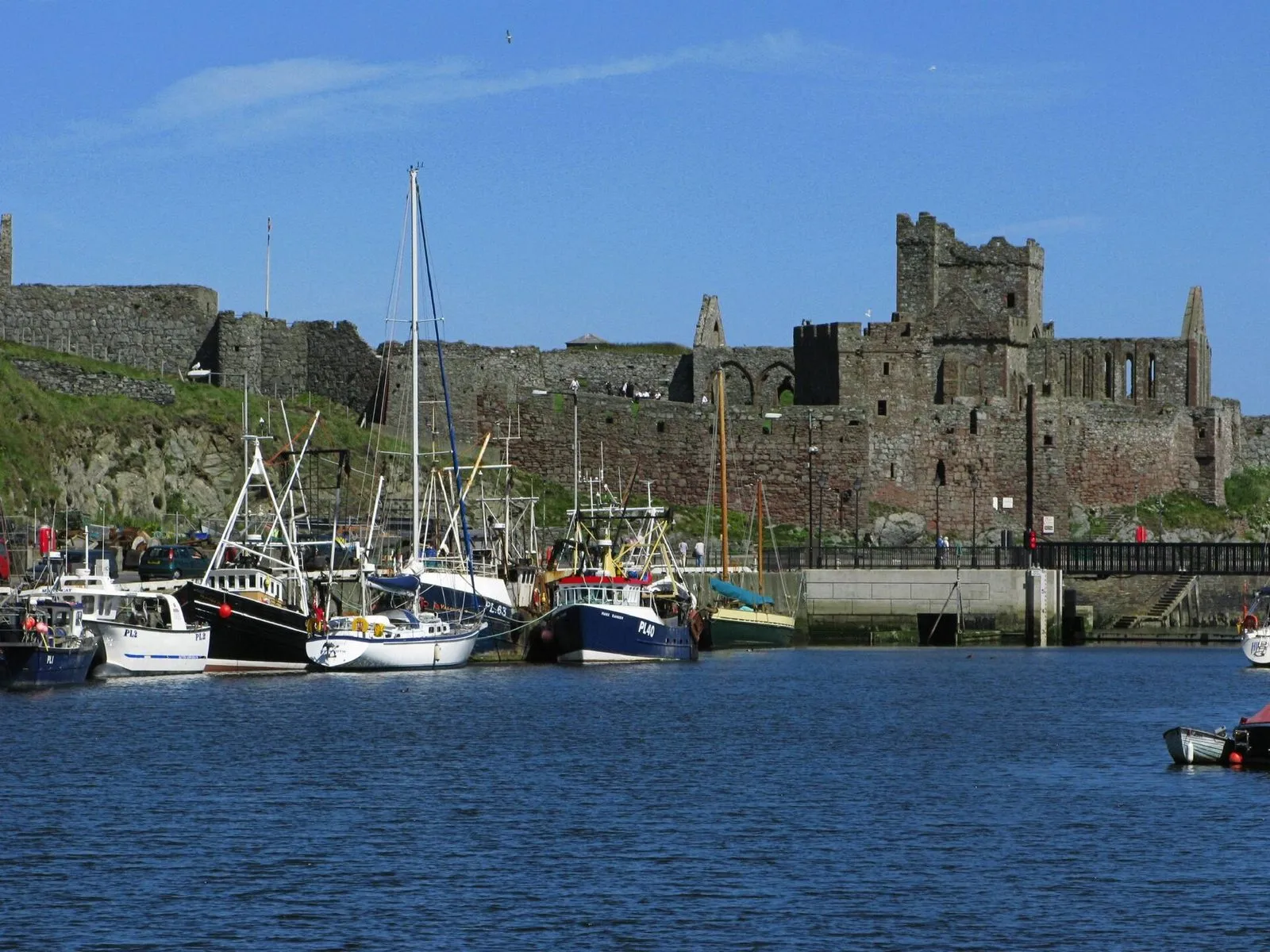 Fishing boats and yachts moored in Peel harbour below the ruined red sandstone walls of Peel Castle, under a blue sky