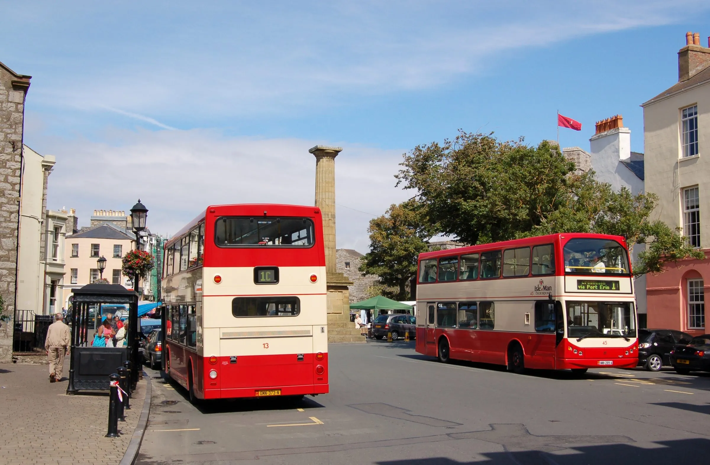 Two red-and-cream double-decker buses in Castletown's Market Square, with the Smelt monument column and Georgian houses behind