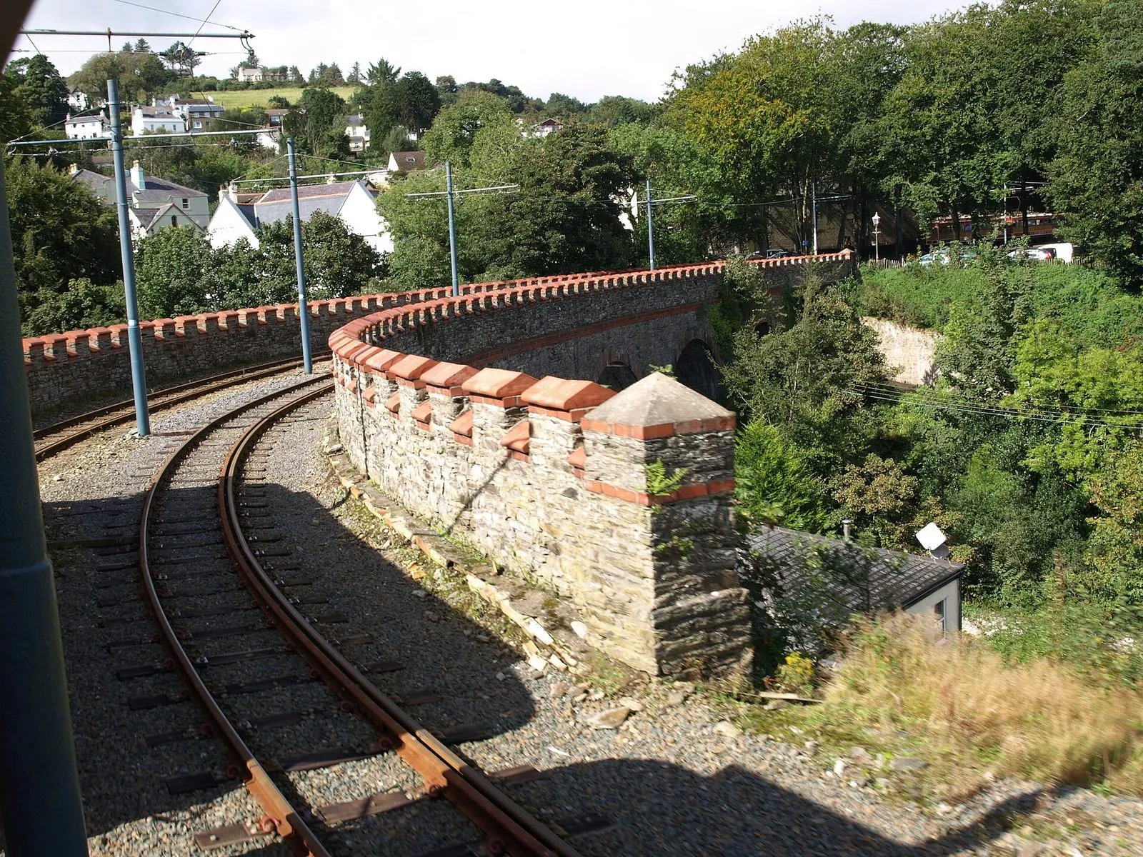 A Manx Electric Railway tram’s view of the curving, castellated stone viaduct into Laxey station, with trees and houses on the hillside