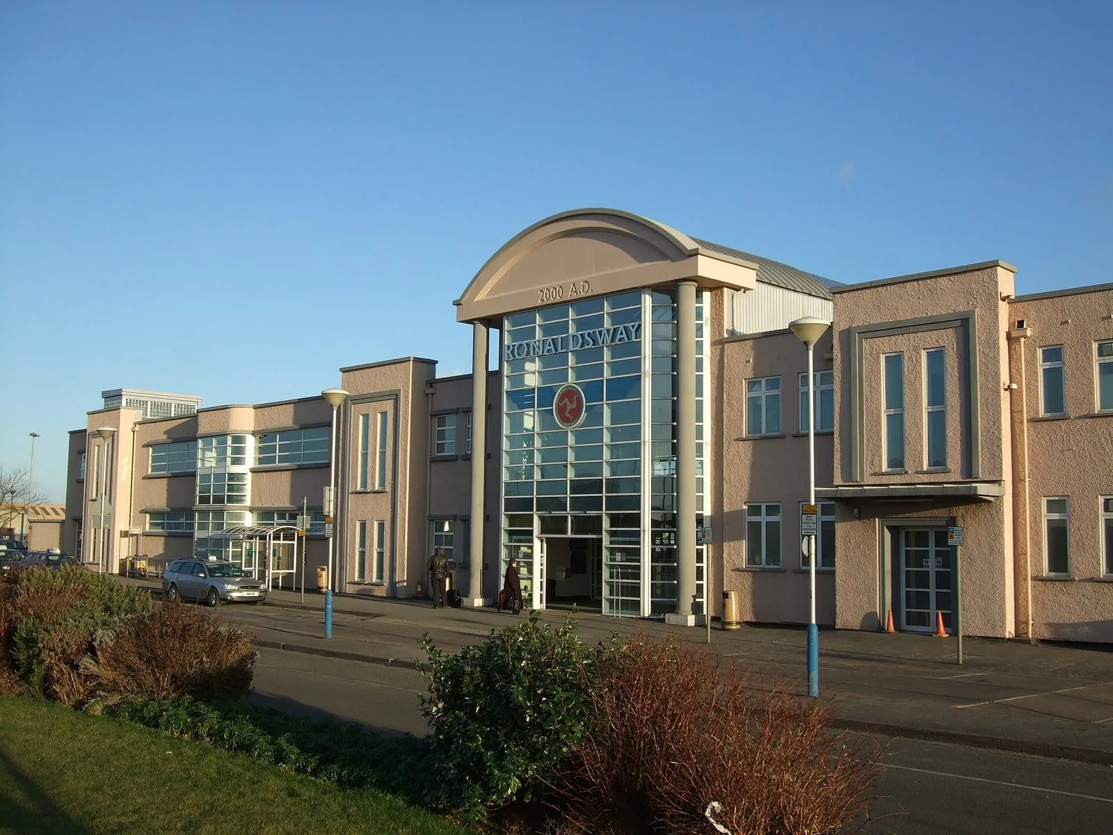 The glass entrance of Ronaldsway airport terminal, with a three-legs emblem and pinkish rendered wings either side, under a clear blue sky