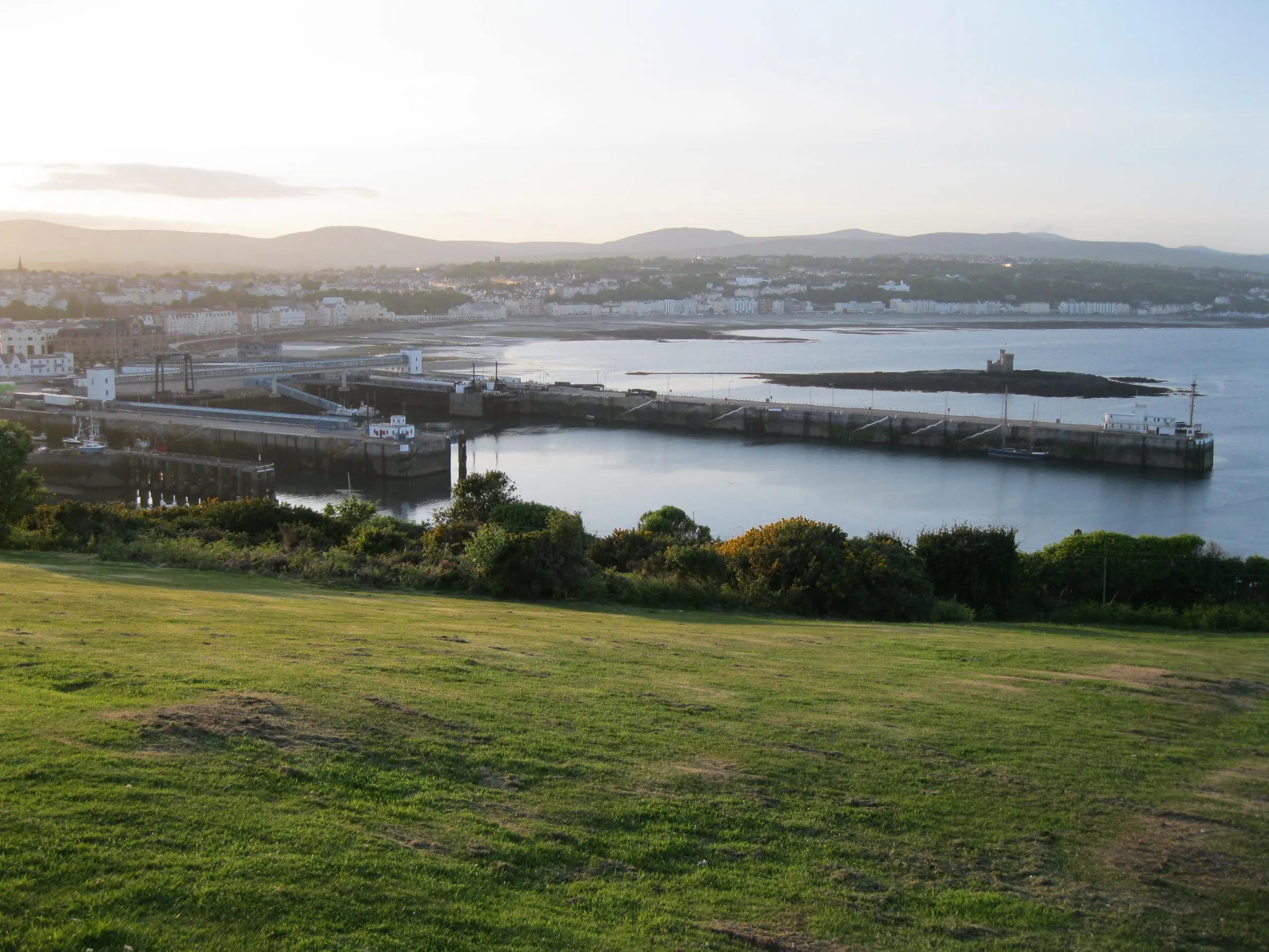 Douglas harbour and its long breakwater seen from Douglas Head in evening light, with the promenade curving round the bay and hills behind