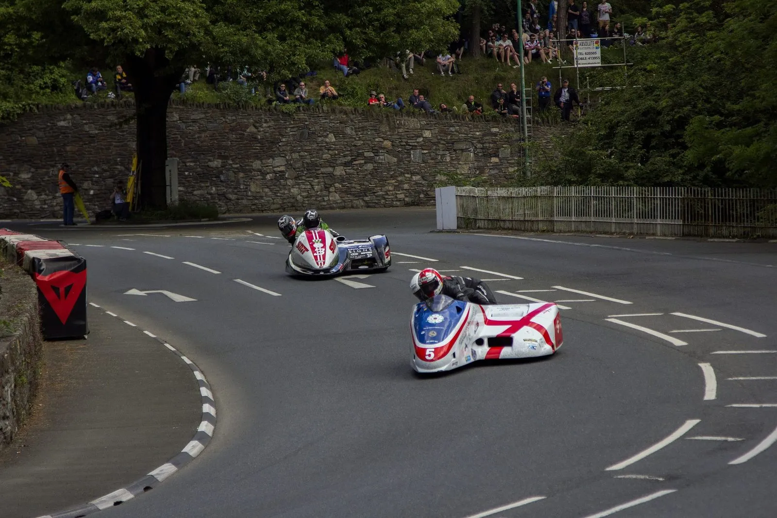 Two sidecar outfits taking the S-bend at Braddan Bridge, with spectators sitting on the bank above the stone wall