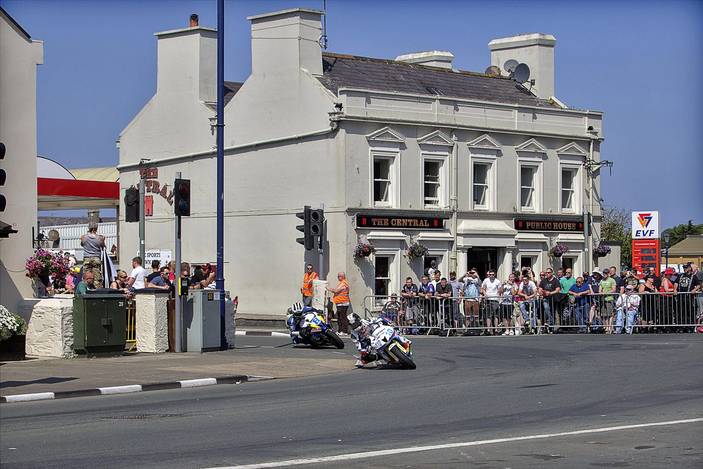 A Supersport rider leaning through Parliament Square in Ramsey past the Central pub, with spectators behind the barriers and marshals in orange
