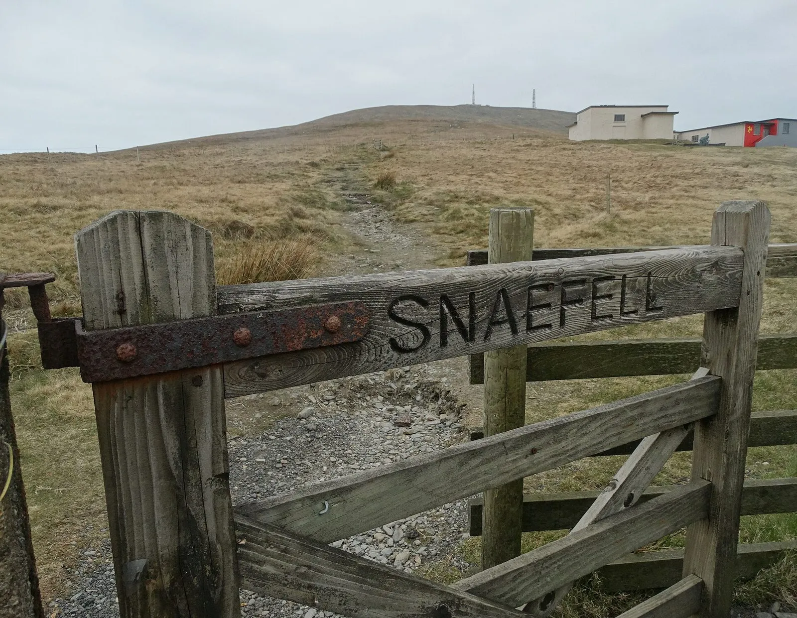 A wooden field gate carved with the word Snaefell, with a stony path climbing a bare grassy hillside behind it towards buildings on the skyline